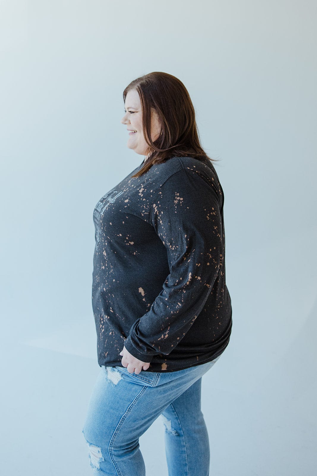 A person stands sideways, wearing the "DOUBLE TROUBLE DRINK AND STUMBLE CLINK THE GLASS TRY NOT TO STUMBLE" graphic tee by Love Marlow, paired with ripped jeans, set against a light gray background.