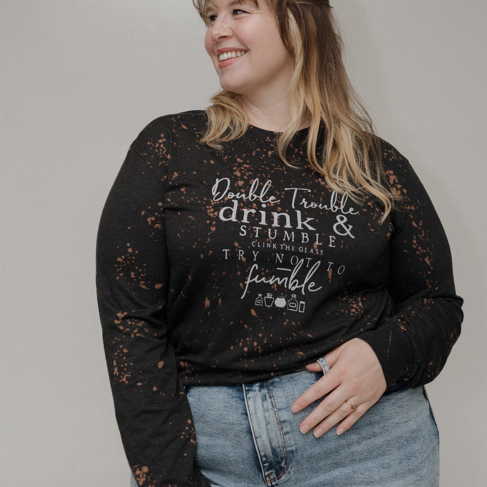"DOUBLE TROUBLE DRINK AND STUMBLE CLINK THE GLASS TRY NOT TO STUMBLE" GRAPHIC TEE Woman wearing a black long-sleeve shirt with text and light blue jeans on a plain background at Love Marlow