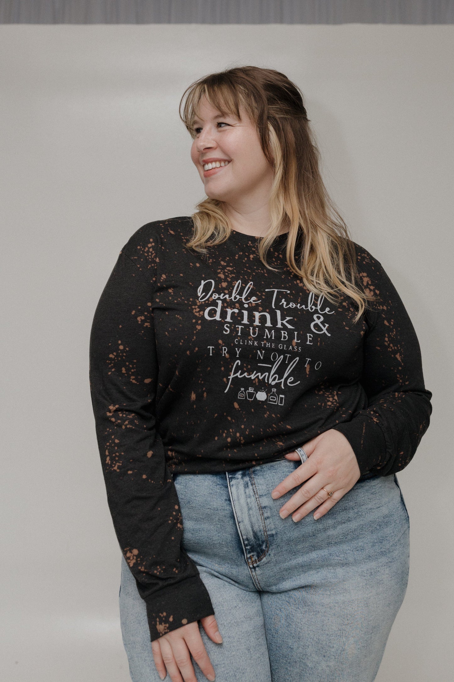 "DOUBLE TROUBLE DRINK AND STUMBLE CLINK THE GLASS TRY NOT TO STUMBLE" GRAPHIC TEE Woman wearing a black long-sleeve shirt with text and light blue jeans on a plain background at Love Marlow