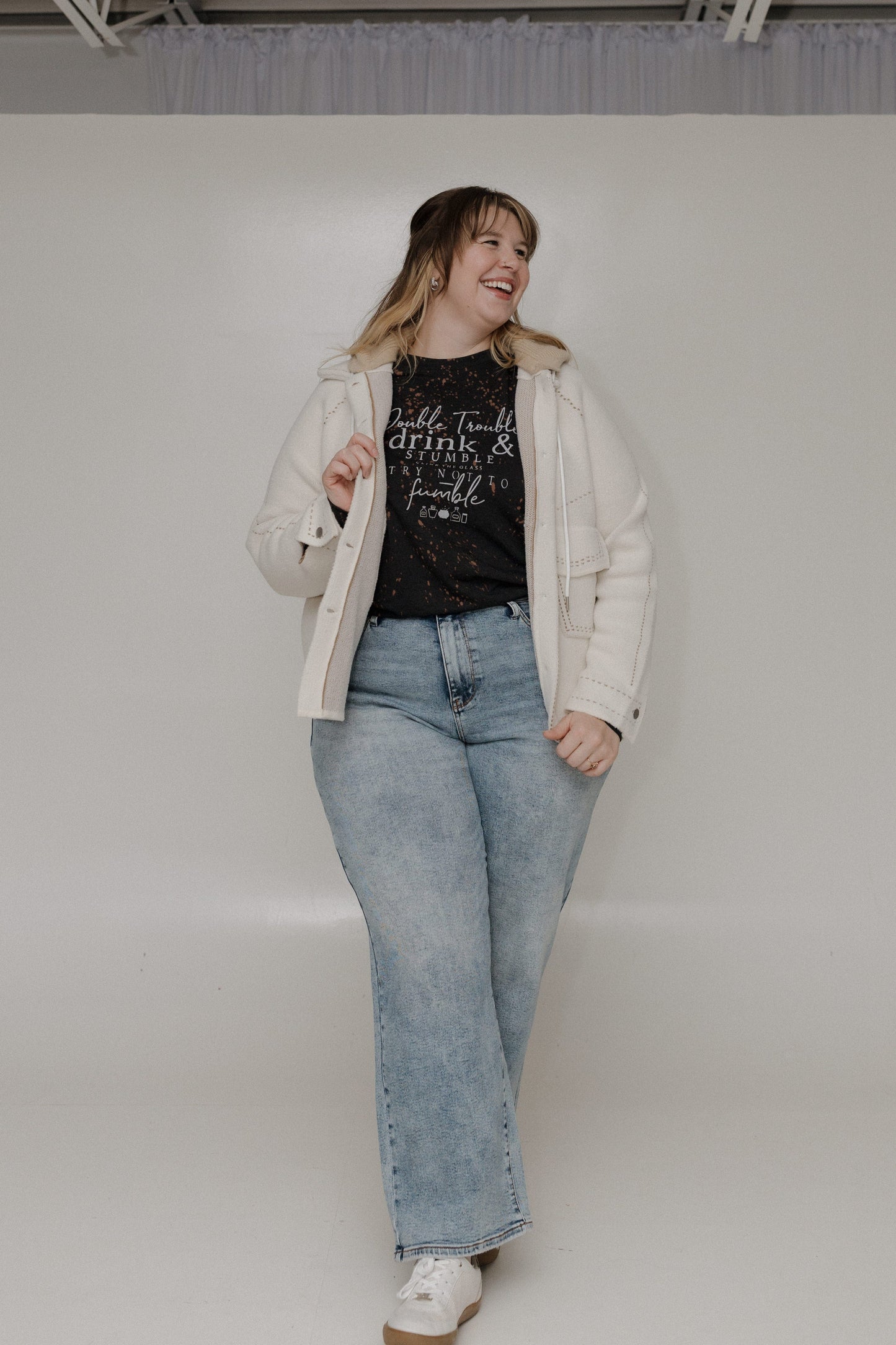 "DOUBLE TROUBLE DRINK AND STUMBLE CLINK THE GLASS TRY NOT TO STUMBLE" GRAPHIC TEE Woman wearing a black long-sleeve shirt with text and light blue jeans on a plain background at Love Marlow