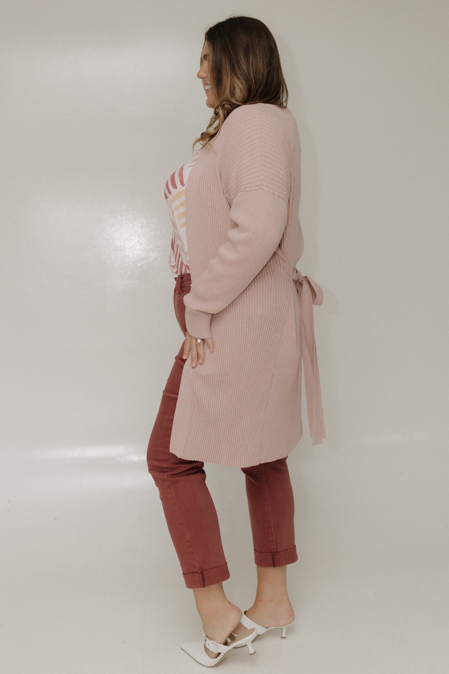 Woman wearing a long pink cardigan and red pants against a white background