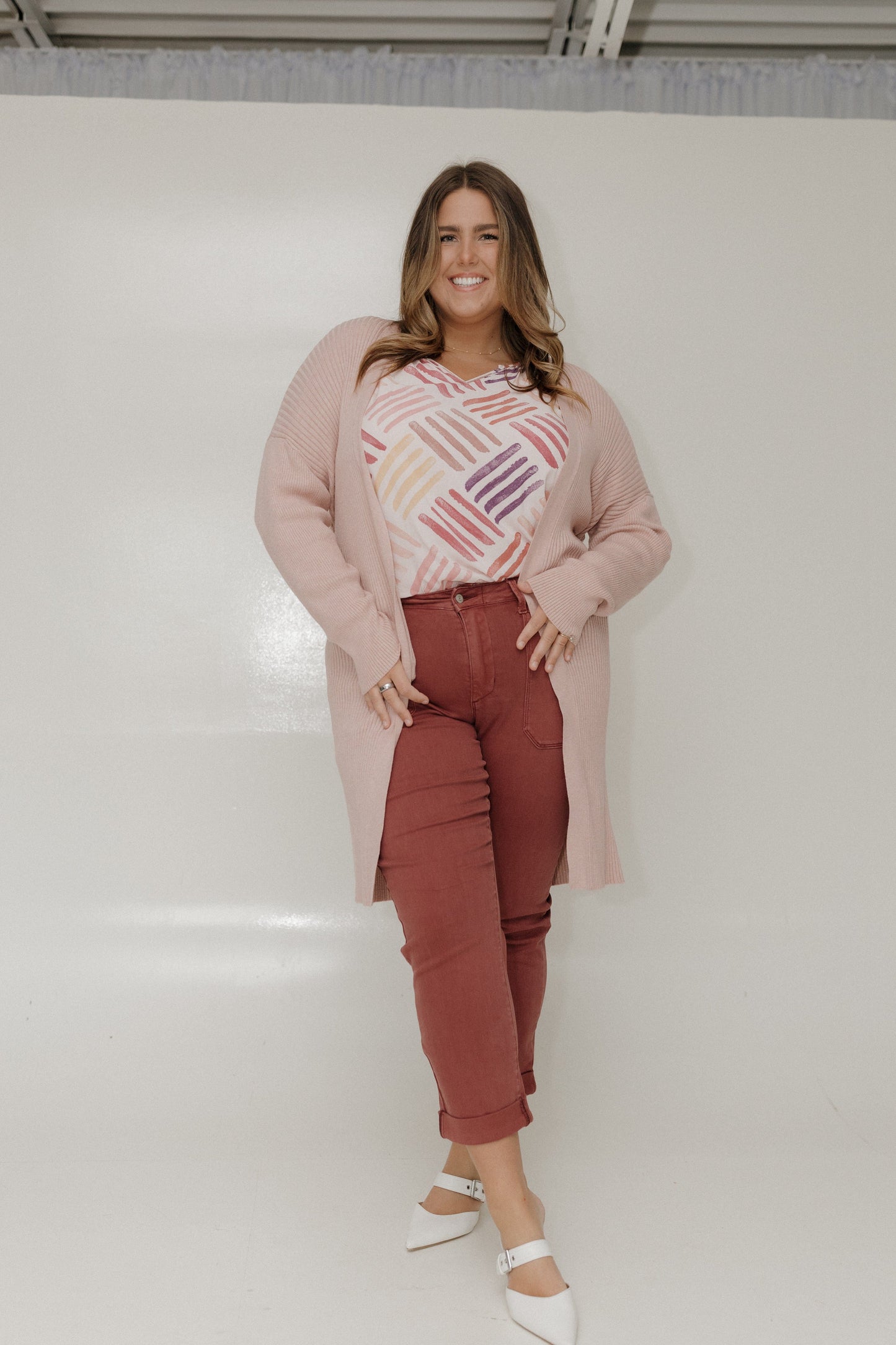 Woman wearing a long pink coat, colorful sweater, and red pants against a white wall.