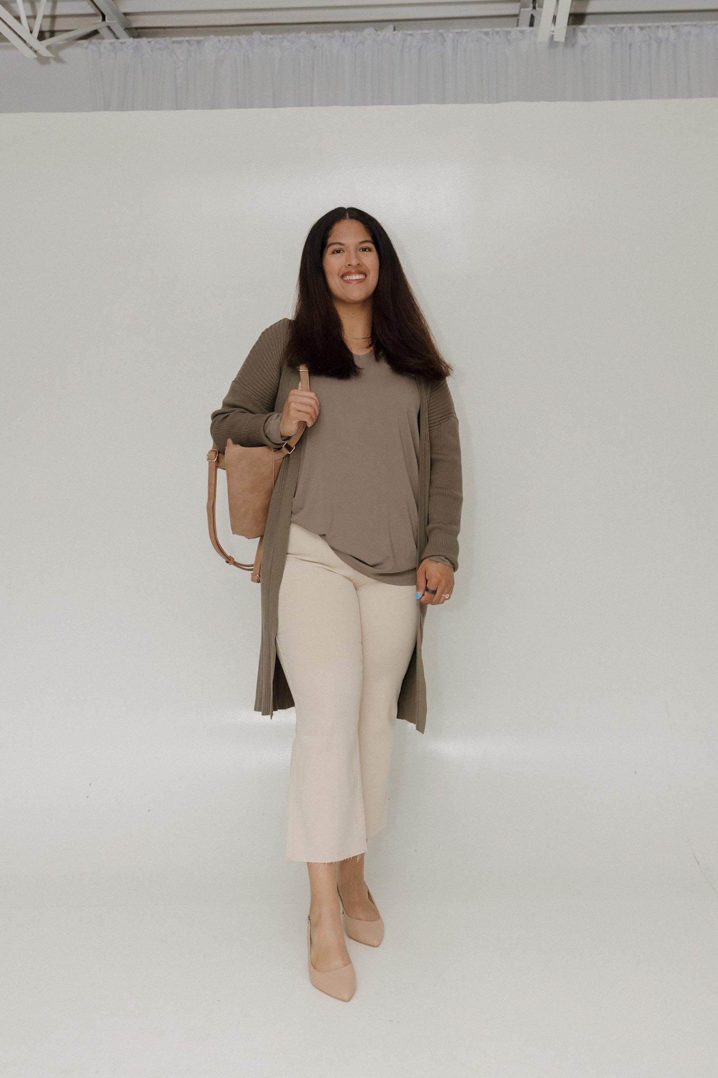 Woman wearing a long beige cardigan, beige top, white pants, and beige shoes against a white background.