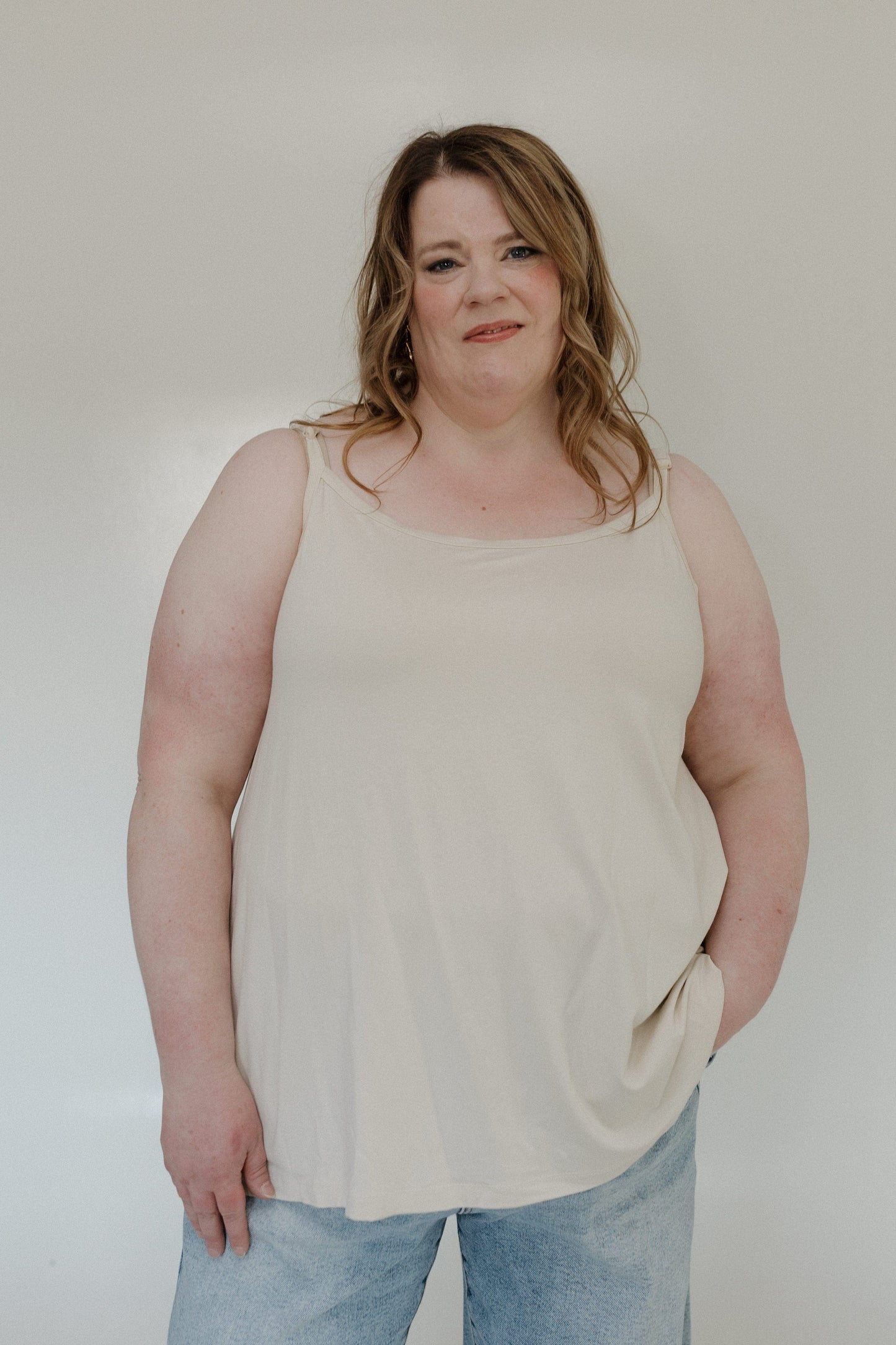 Woman wearing a beige tank top and light blue jeans against a plain background