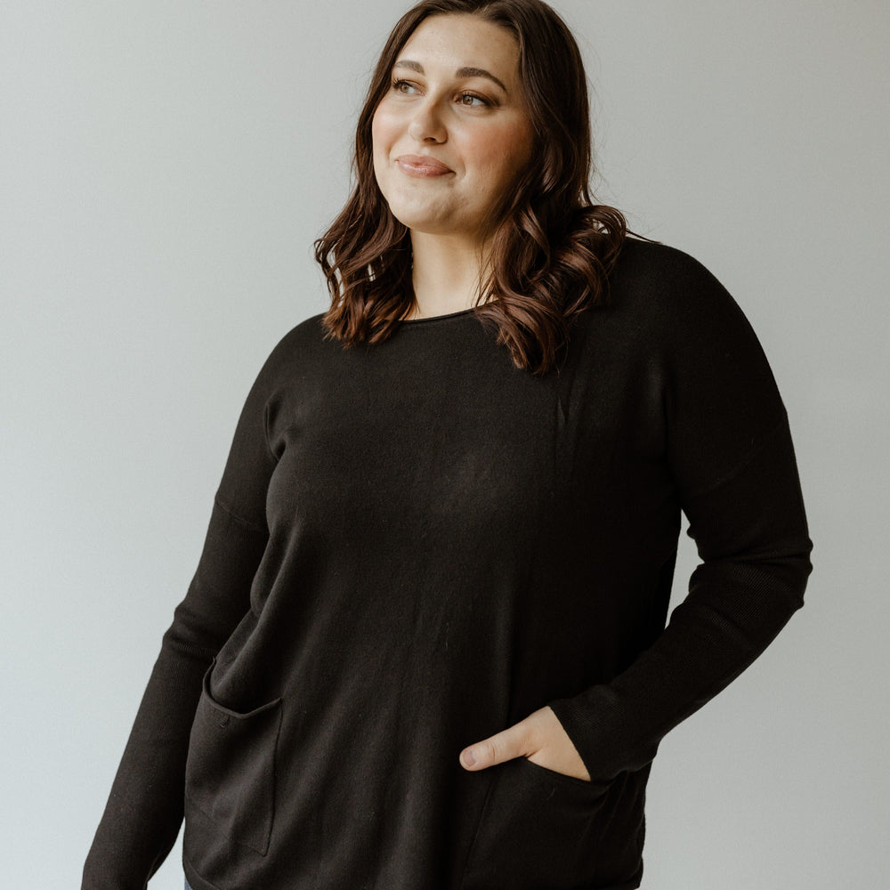 A person with long hair wearing a black crew neck sweater and blue jeans stands against a plain background.