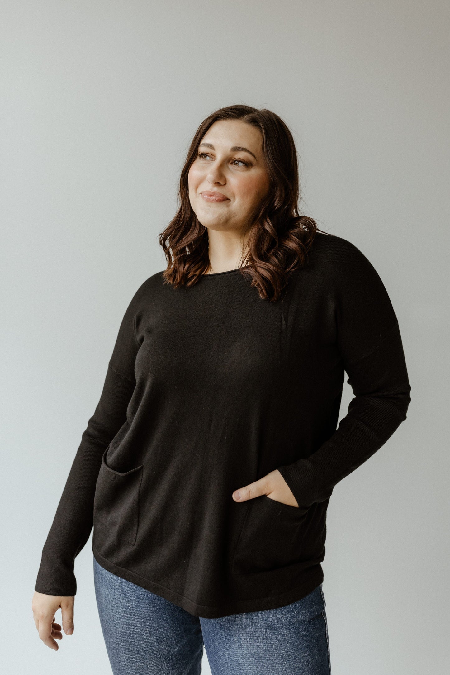 A person with long hair wearing a black crew neck sweater and blue jeans stands against a plain background.