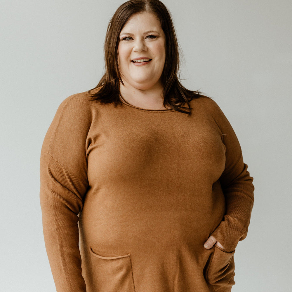 A person with shoulder-length brown hair smiles, donning a slim fit crew neck sweater in Urban Bronze paired with blue jeans, standing against a plain backdrop.