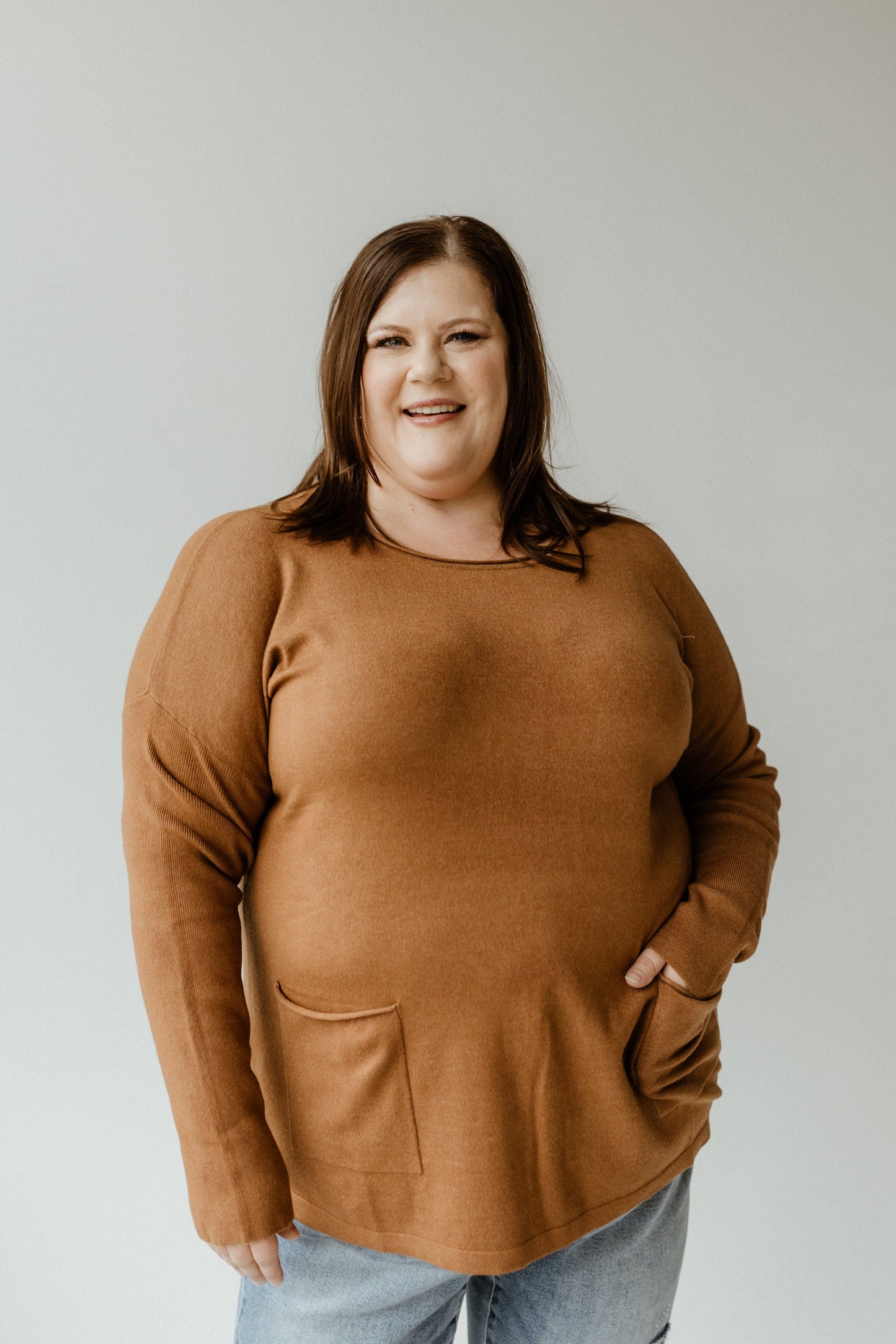 A person with shoulder-length brown hair smiles, donning a slim fit crew neck sweater in Urban Bronze paired with blue jeans, standing against a plain backdrop.