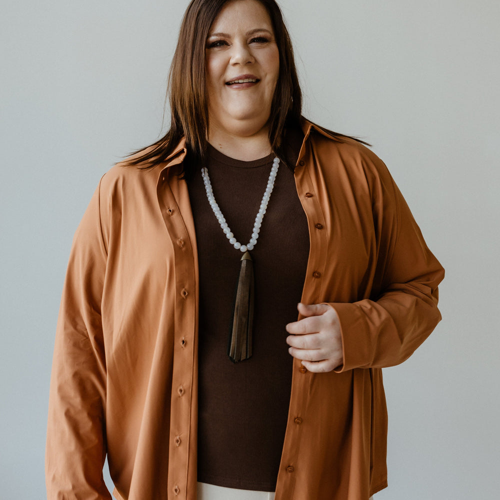 A person wearing a brown Essential Lightweight Button-Up, roasted cinnamon overshirt, and white pants stands smiling against a plain background.