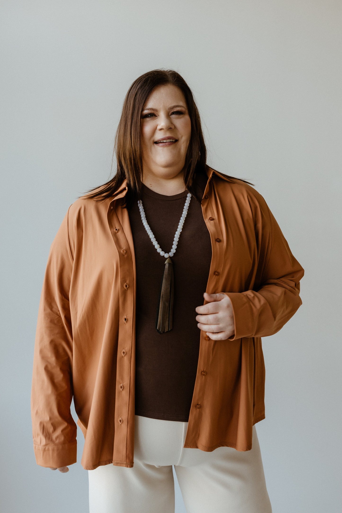 A person wearing a brown Essential Lightweight Button-Up, roasted cinnamon overshirt, and white pants stands smiling against a plain background.