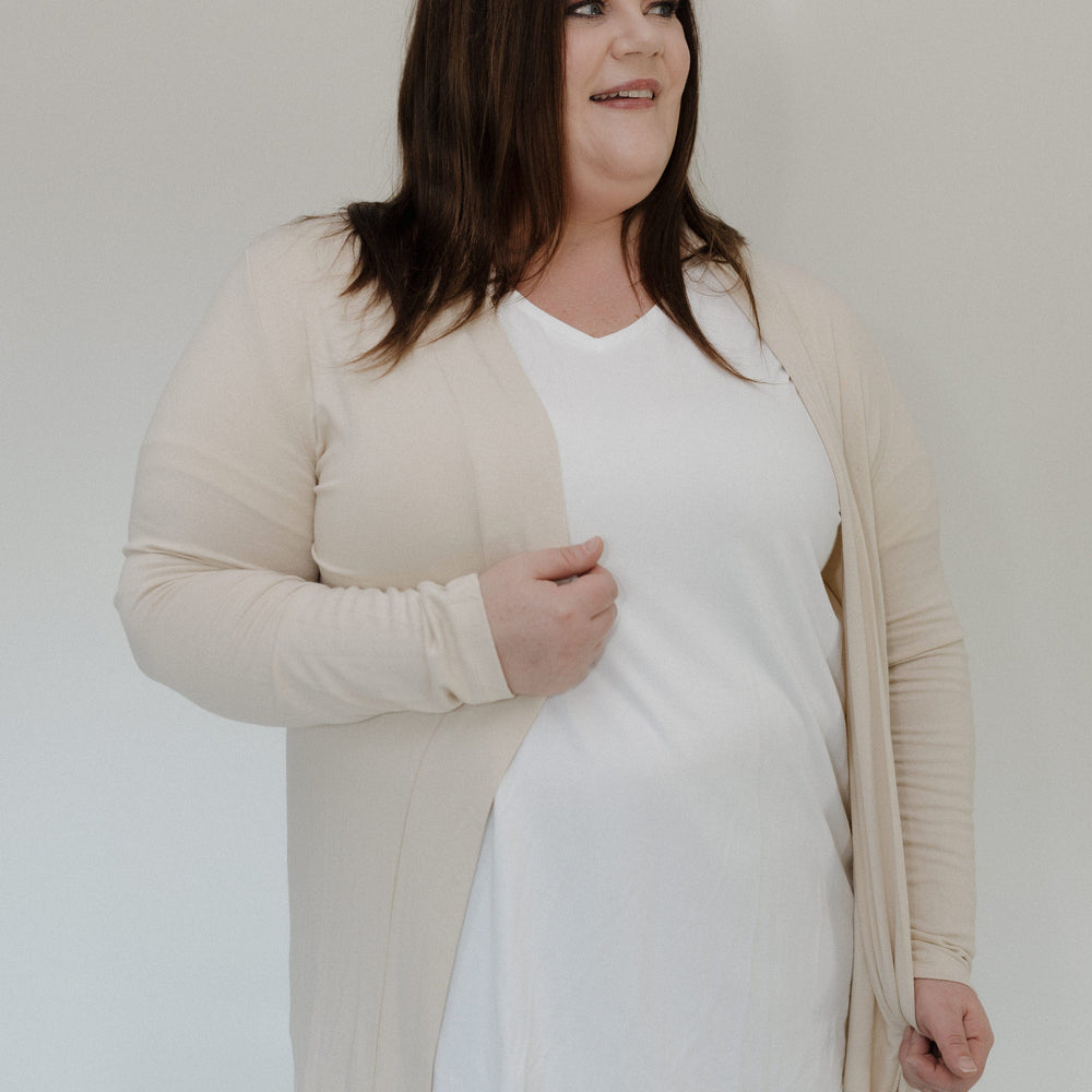 Woman wearing a beige cardigan over a white top and gray pants on a plain background