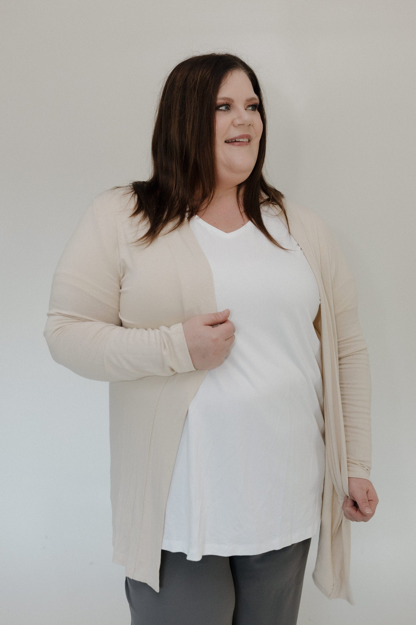 Woman wearing a beige cardigan over a white top and gray pants on a plain background