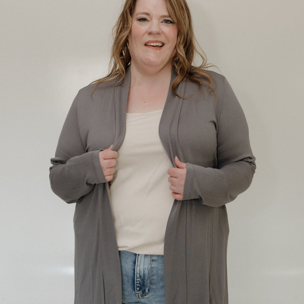 Woman wearing a gray cardigan over a white shirt and blue jeans against a plain background