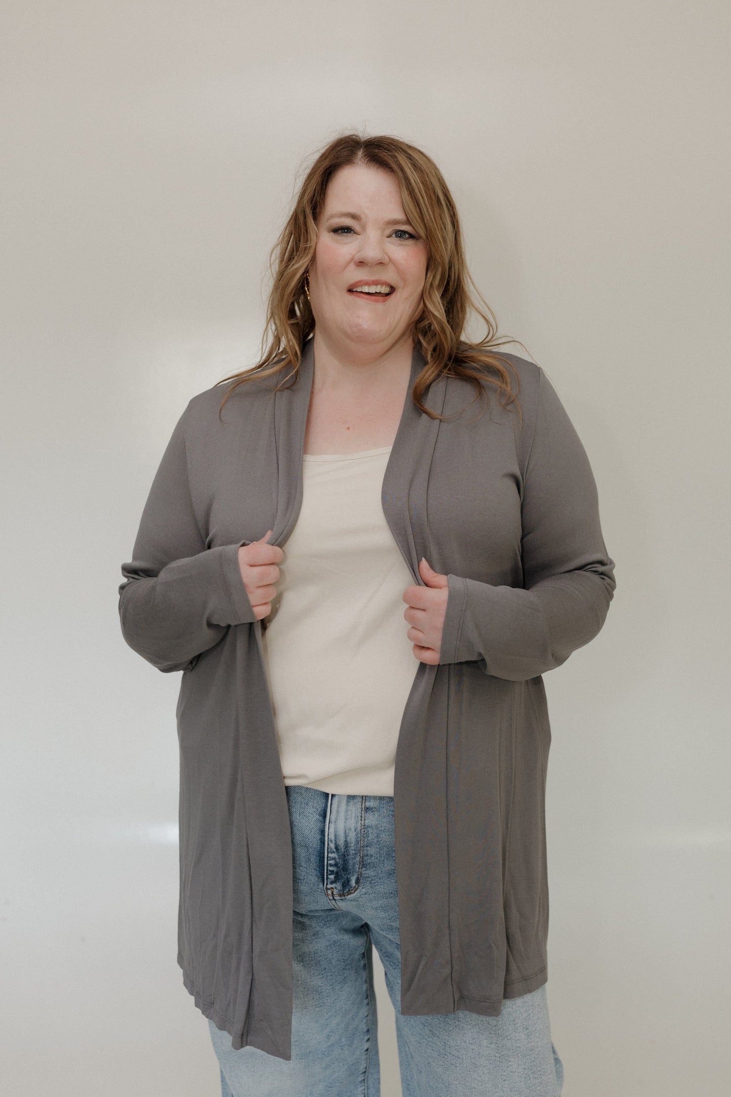 Woman wearing a gray cardigan over a white shirt and blue jeans against a plain background