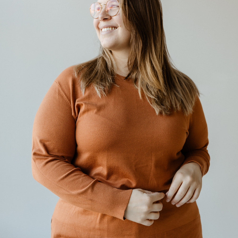 A person wearing glasses and a spiced rose, lightweight tunic sweater stands smiling with hands relaxed in front, set against a plain background.