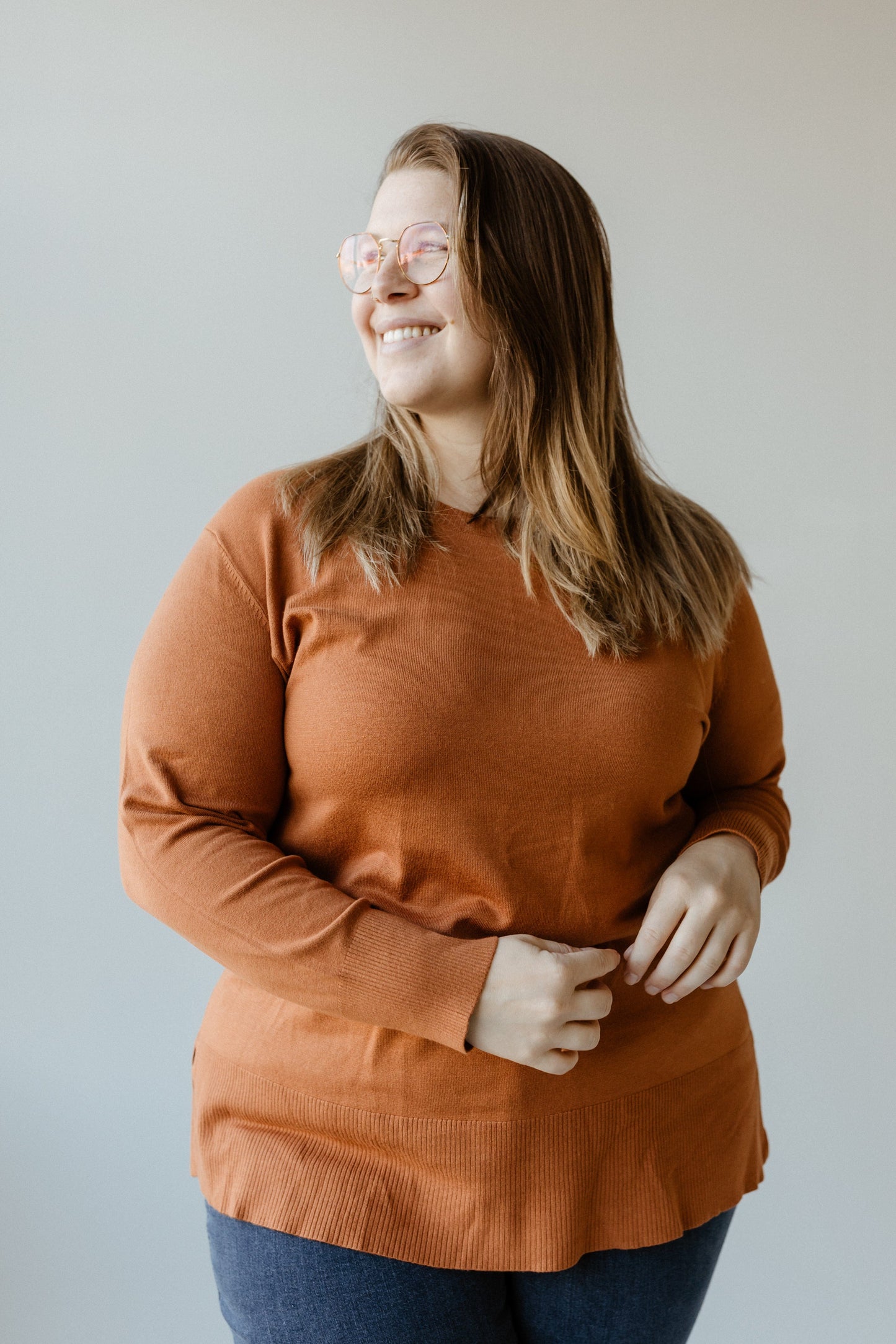 A person wearing glasses and a spiced rose, lightweight tunic sweater stands smiling with hands relaxed in front, set against a plain background.