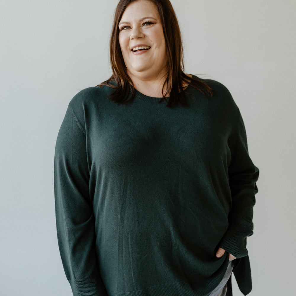 A person in a sophisticated, essential lightweight tunic sweater and ripped jeans stands against a plain backdrop, smiling.