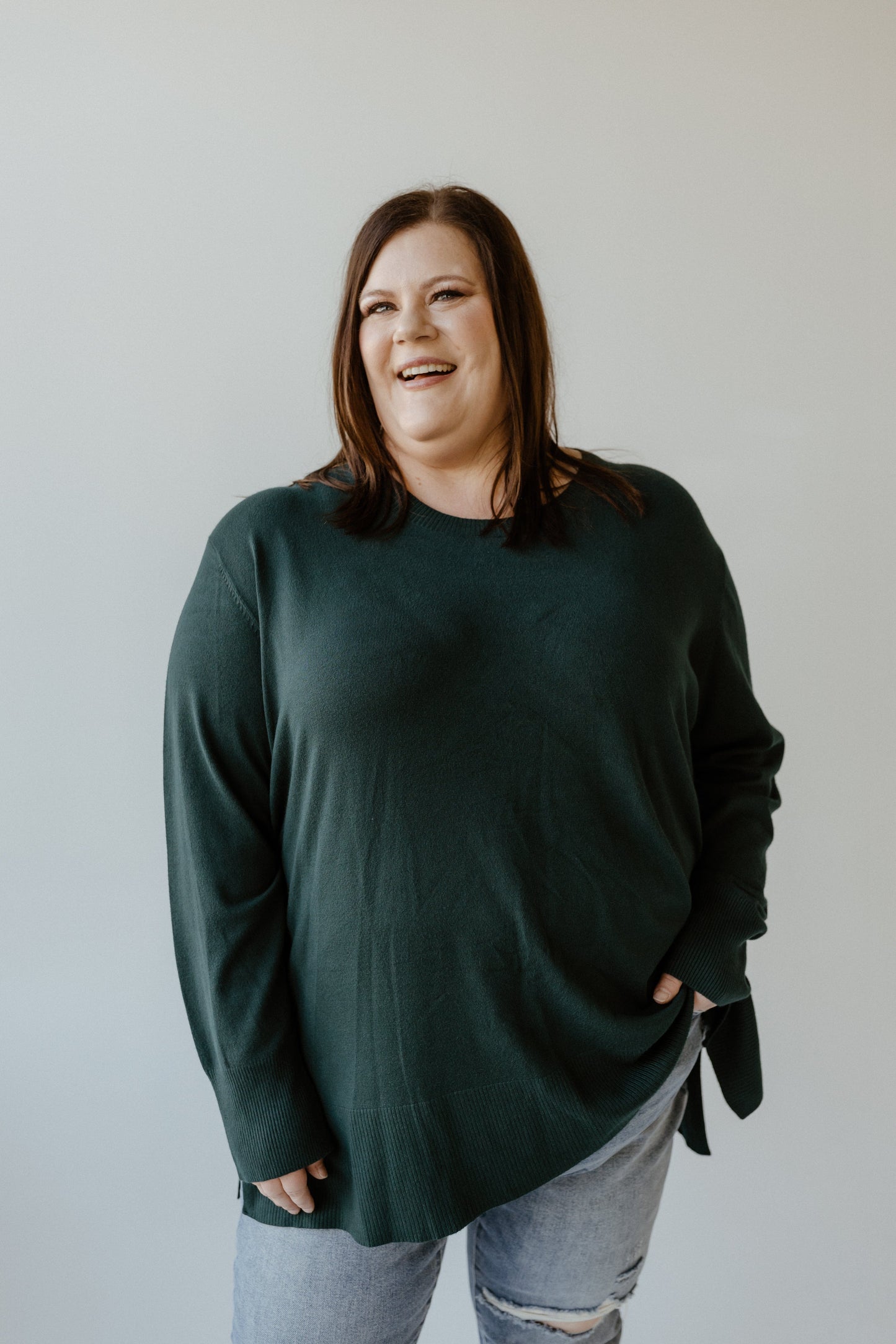 A person in a sophisticated, essential lightweight tunic sweater and ripped jeans stands against a plain backdrop, smiling.