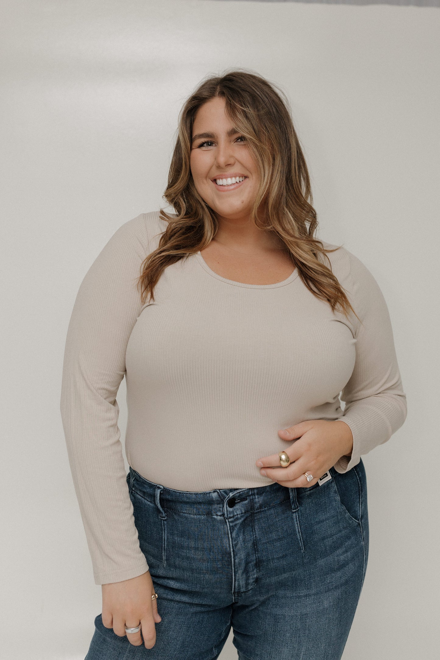 Woman wearing a beige long-sleeve top and blue jeans against a plain background