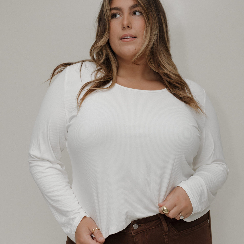Woman wearing a white long-sleeve top and brown pants against a plain background