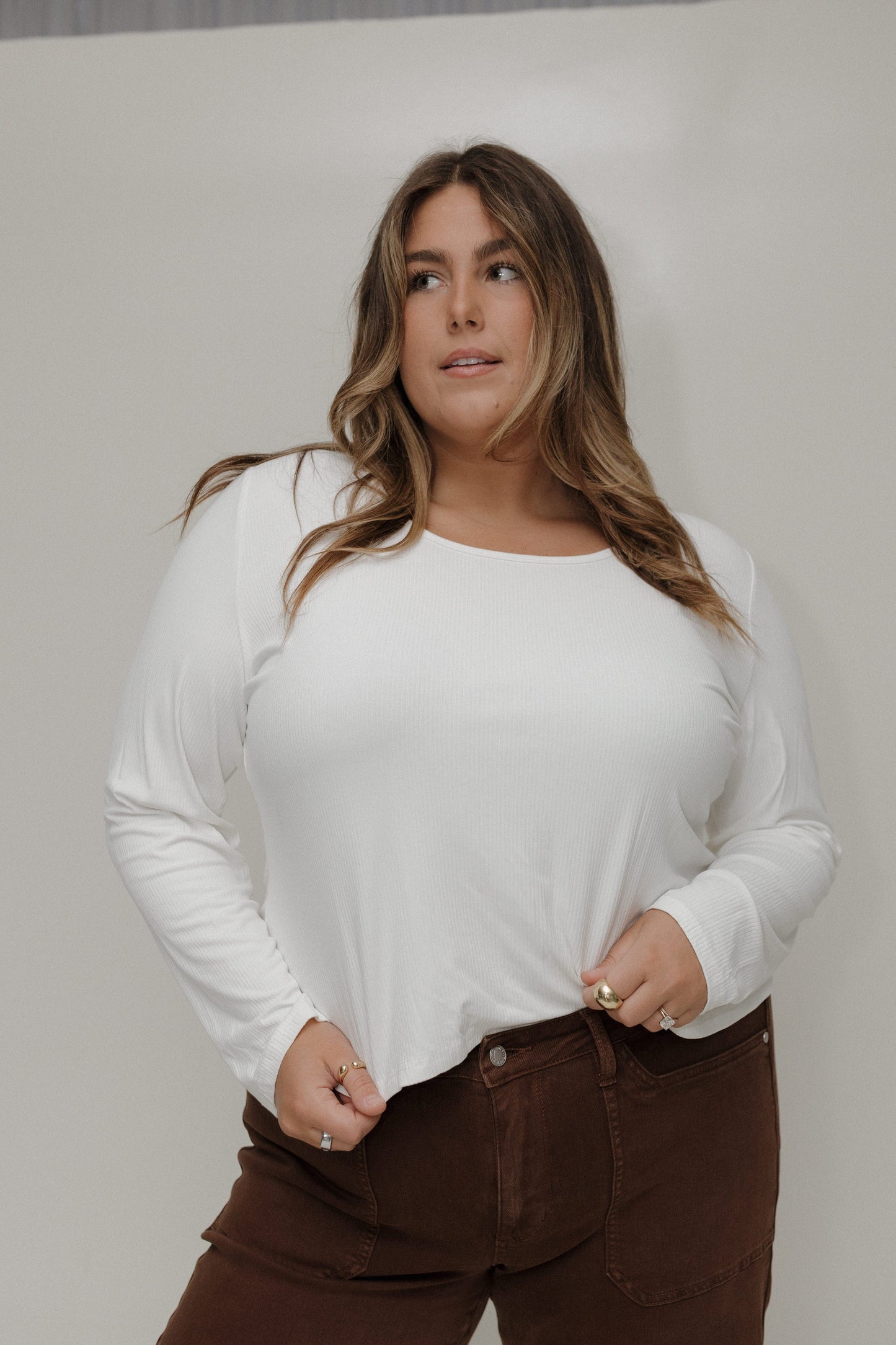 Woman wearing a white long-sleeve top and brown pants against a plain background