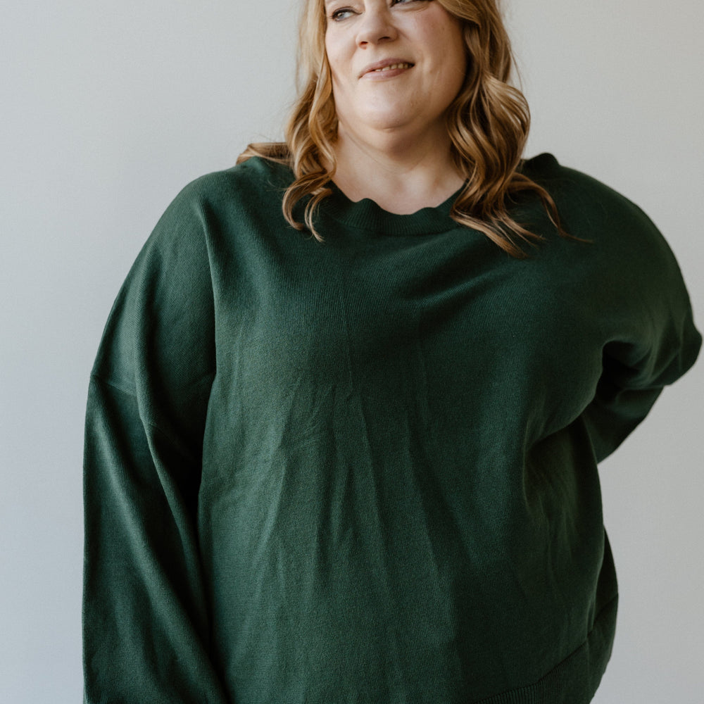 A person with long hair, wearing an Essential Sweater in dark green, stands against a light background, looking to the side.