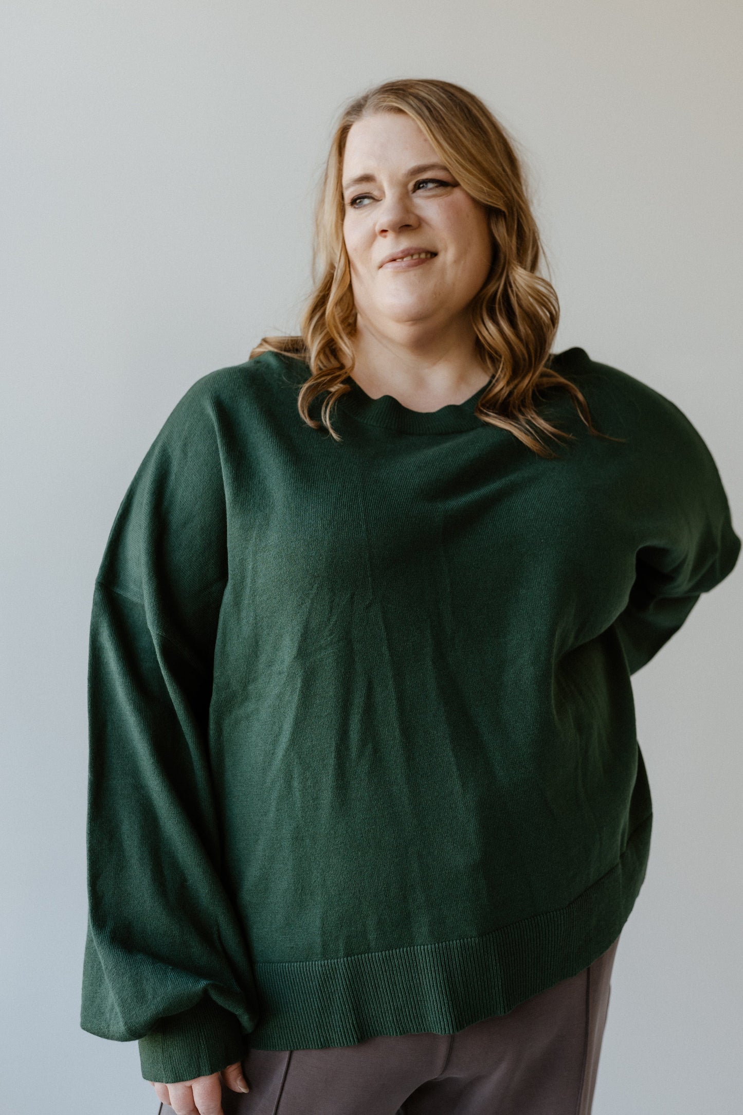 A person with long hair, wearing an Essential Sweater in dark green, stands against a light background, looking to the side.