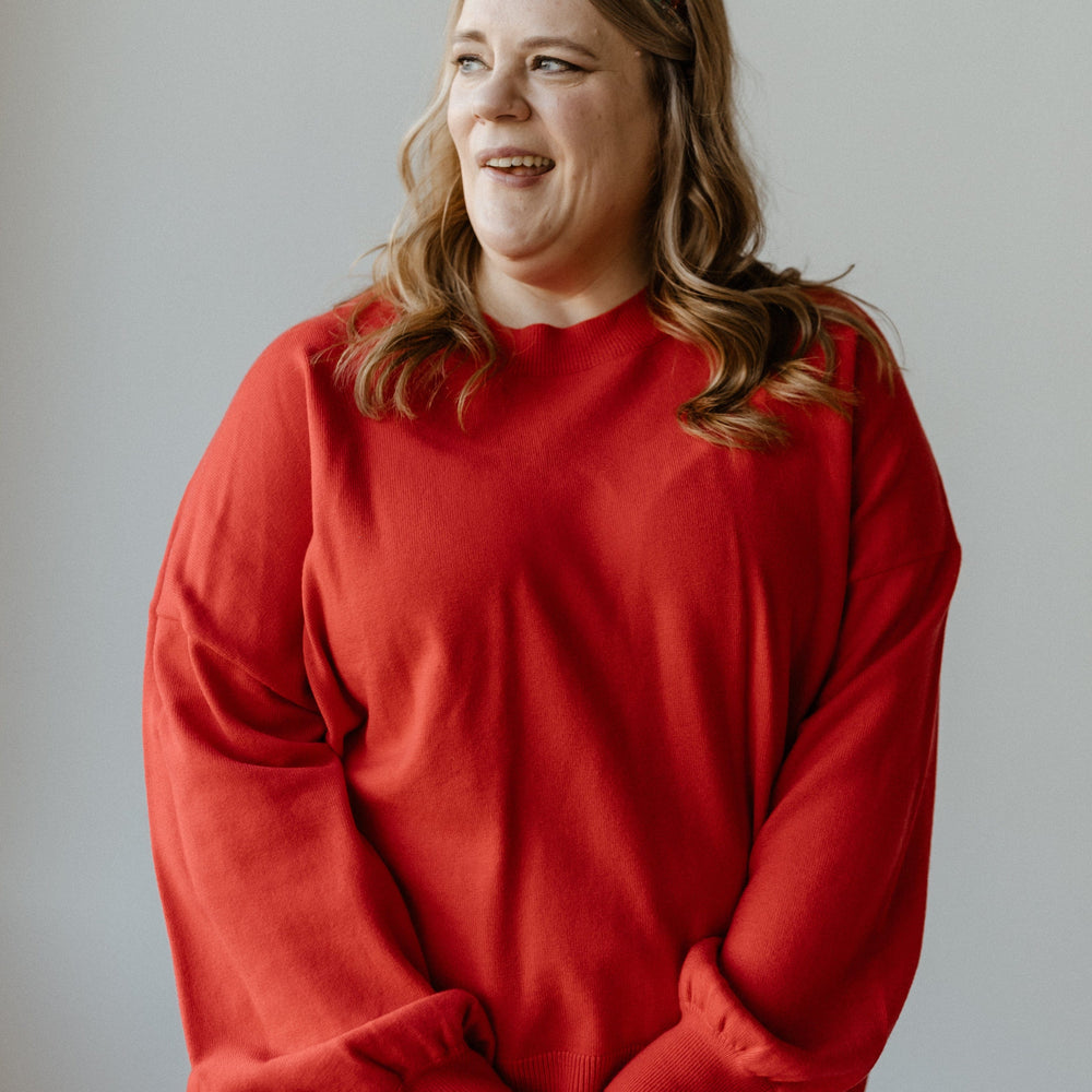 A person wearing a bold red sweater and headband with loose curls stands against a plain background, looking to the right.