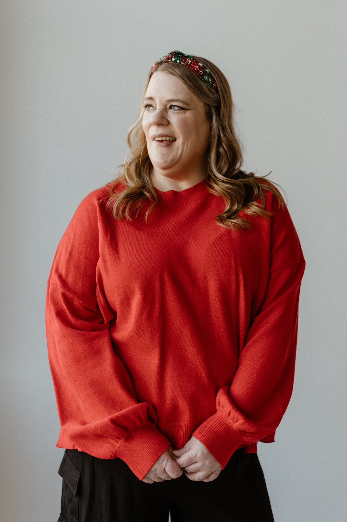 A person wearing a bold red sweater and headband with loose curls stands against a plain background, looking to the right.