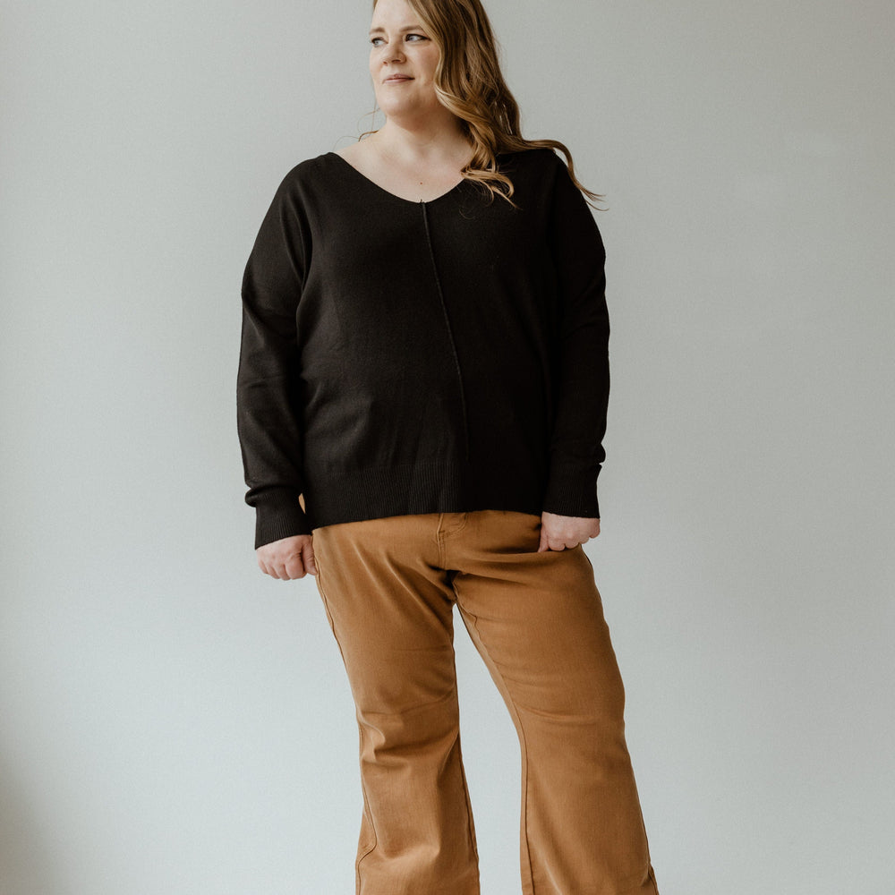 A person wearing an Essential black sweater and brown pants stands against a plain light background.