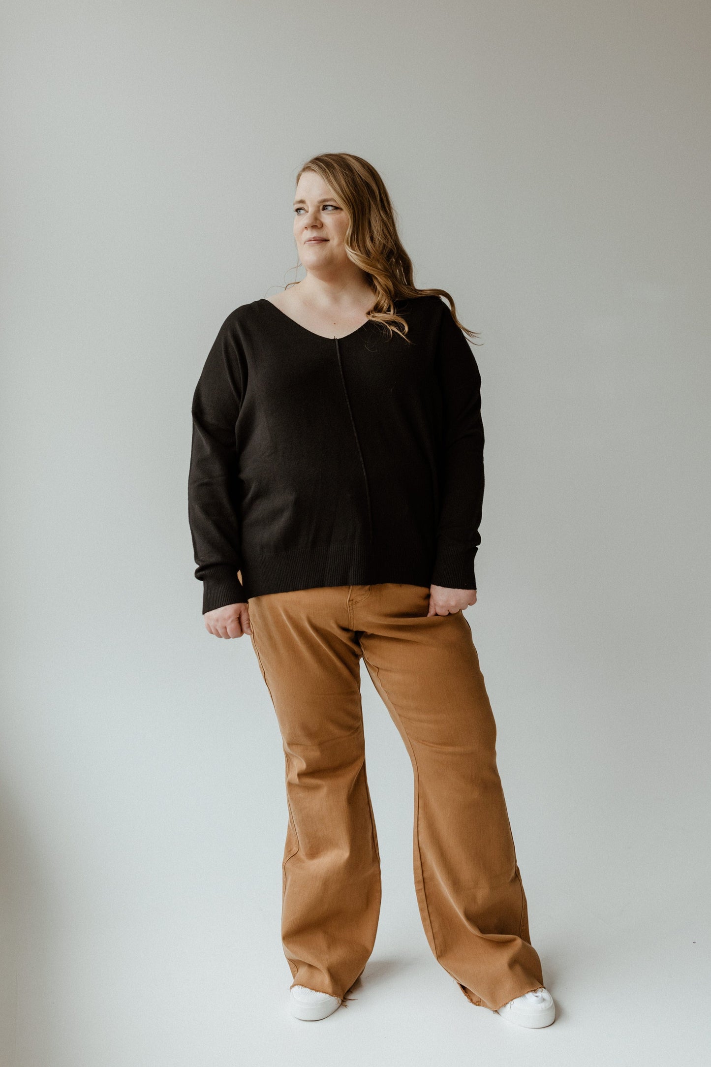 A person wearing an Essential black sweater and brown pants stands against a plain light background.