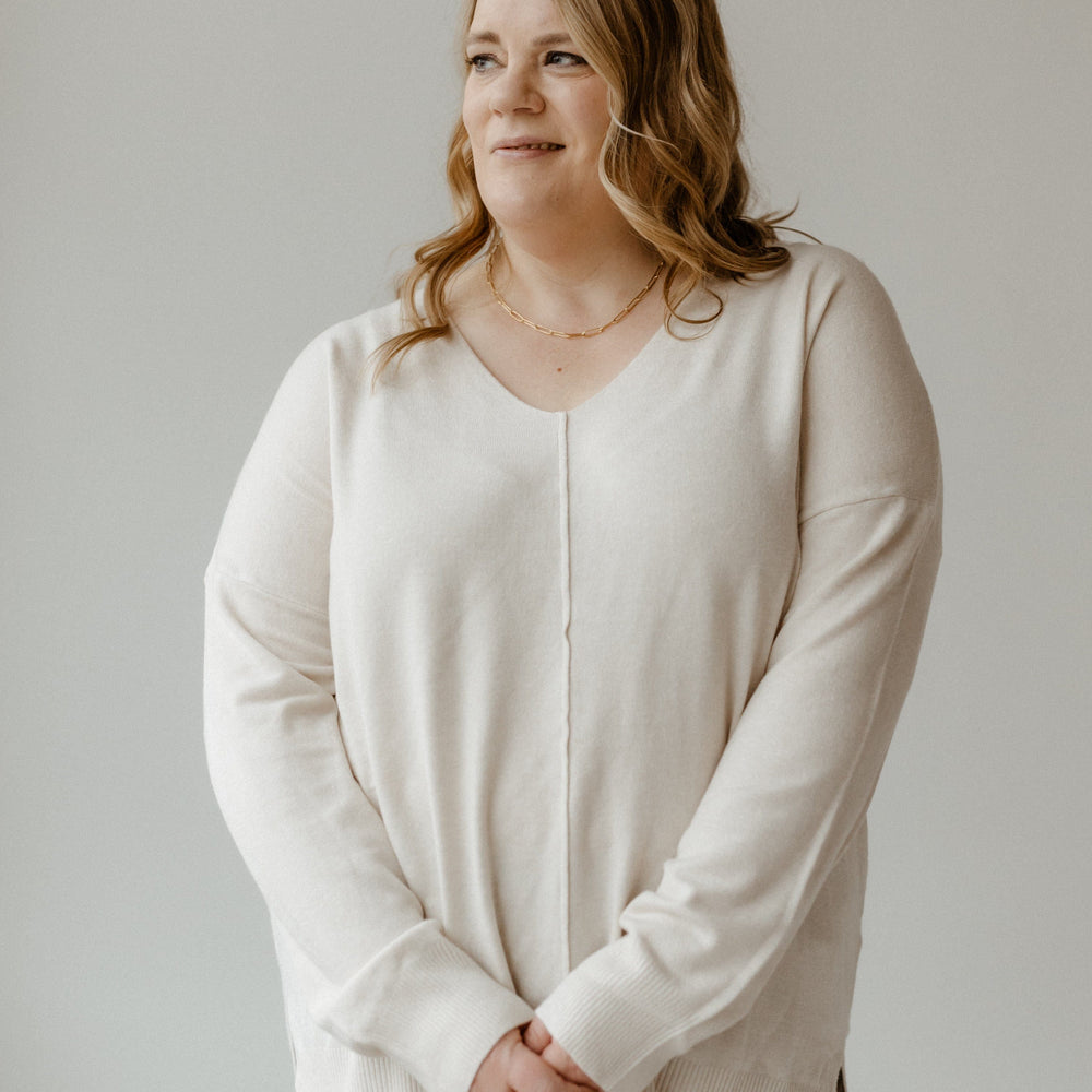 A person with long hair stands against a plain background, wearing a light, slim fit sweater featuring a V-neck and front seam details, paired with gray pants, looking to the side.