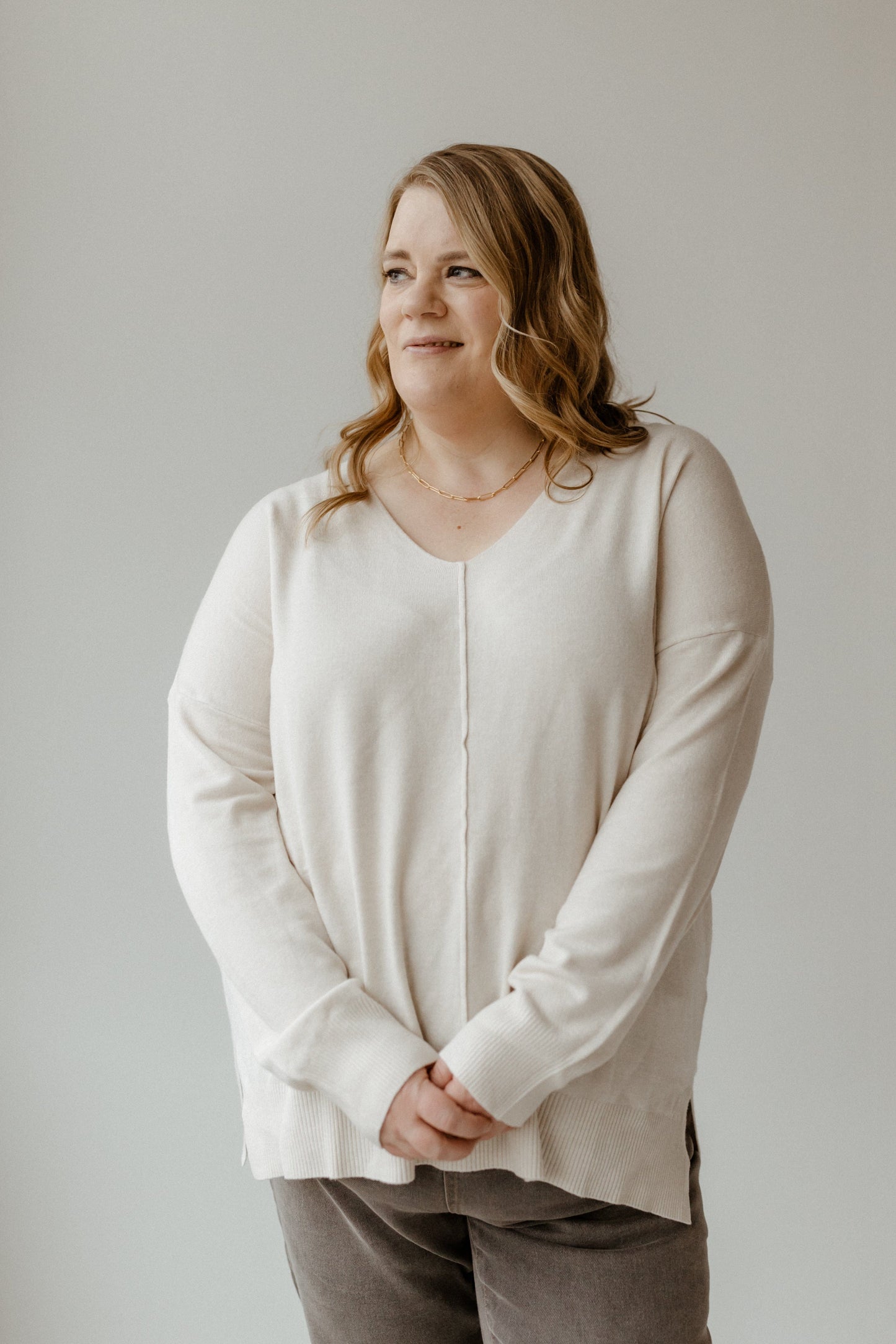 A person with long hair stands against a plain background, wearing a light, slim fit sweater featuring a V-neck and front seam details, paired with gray pants, looking to the side.