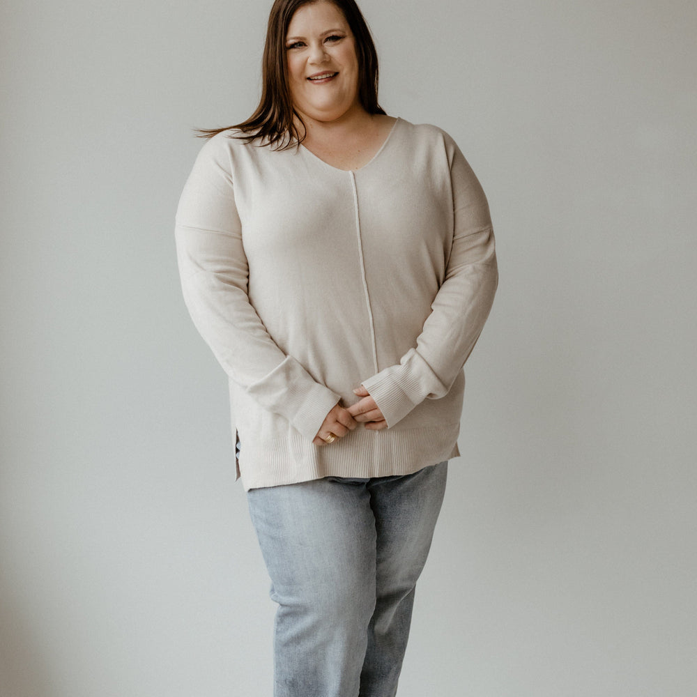 A person wearing a cozy V-Neck Front Seam Sweater and jeans stands against a plain background, smiling with hands clasped in front.