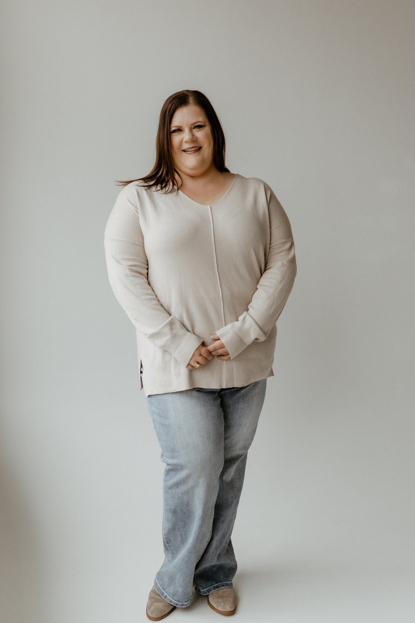 A person wearing a cozy V-Neck Front Seam Sweater and jeans stands against a plain background, smiling with hands clasped in front.