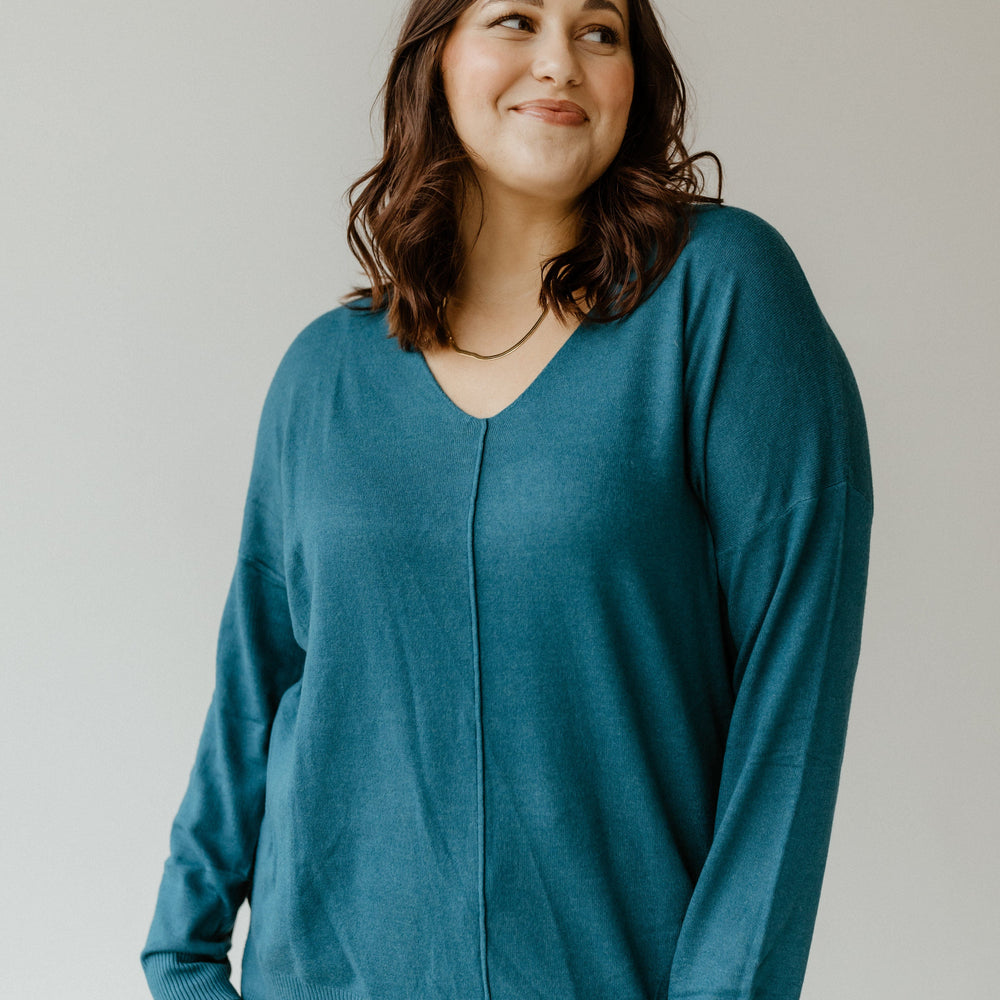 A person with shoulder-length dark hair wearing a teal front seam sweater stands against a plain background and looks slightly to the side.