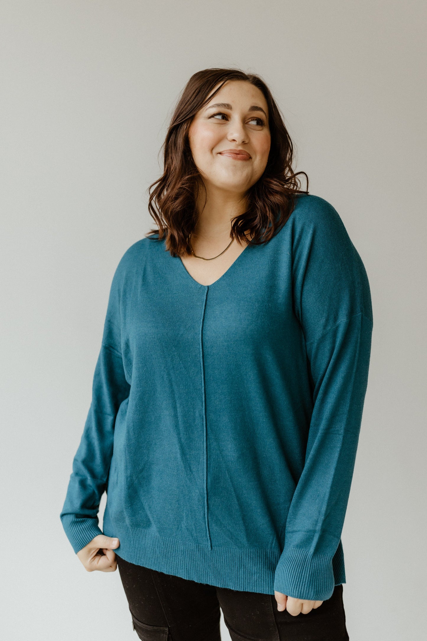 A person with shoulder-length dark hair wearing a teal front seam sweater stands against a plain background and looks slightly to the side.