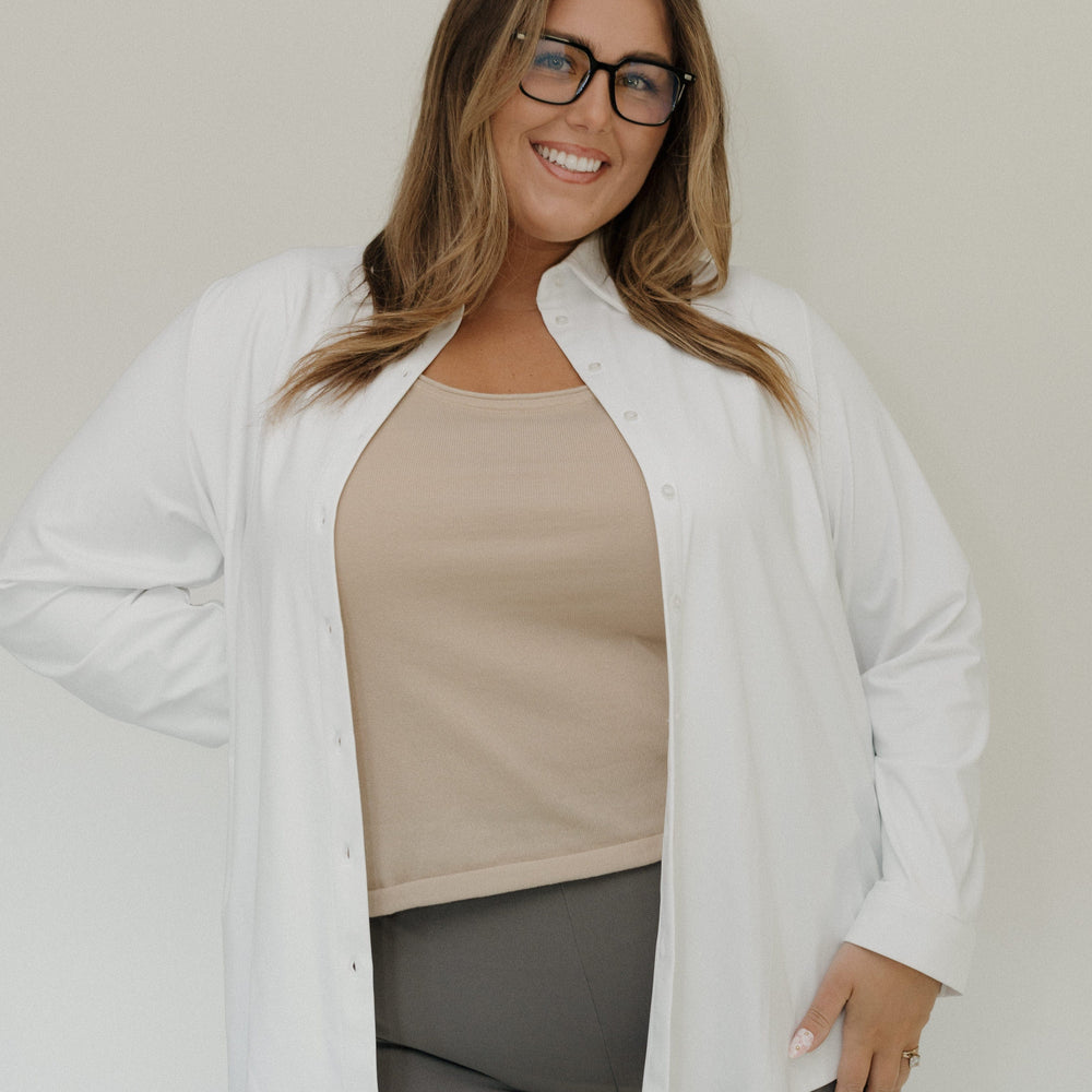 Woman wearing a white cardigan over a beige top and gray pants against a plain background
