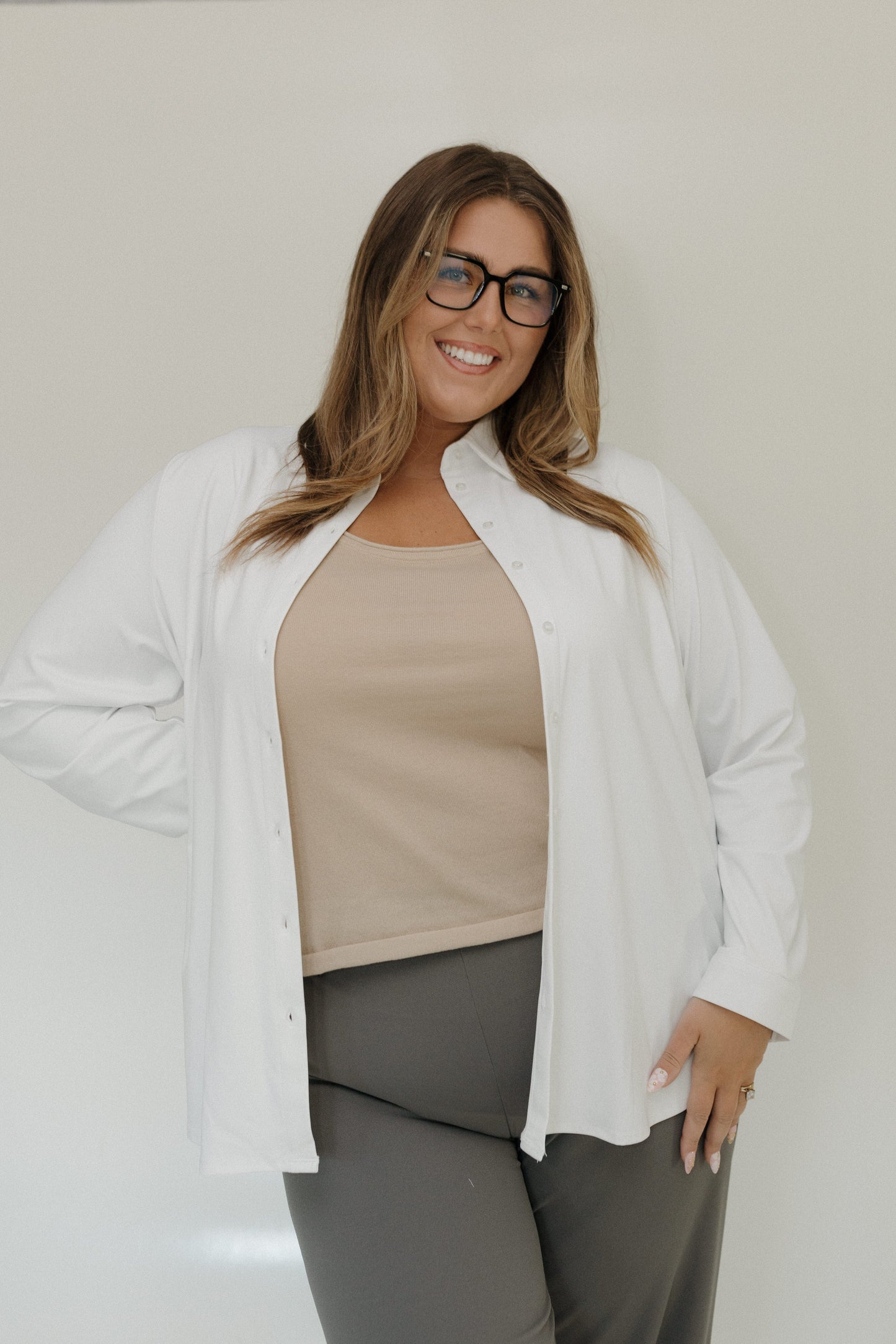 Woman wearing a white cardigan over a beige top and gray pants against a plain background