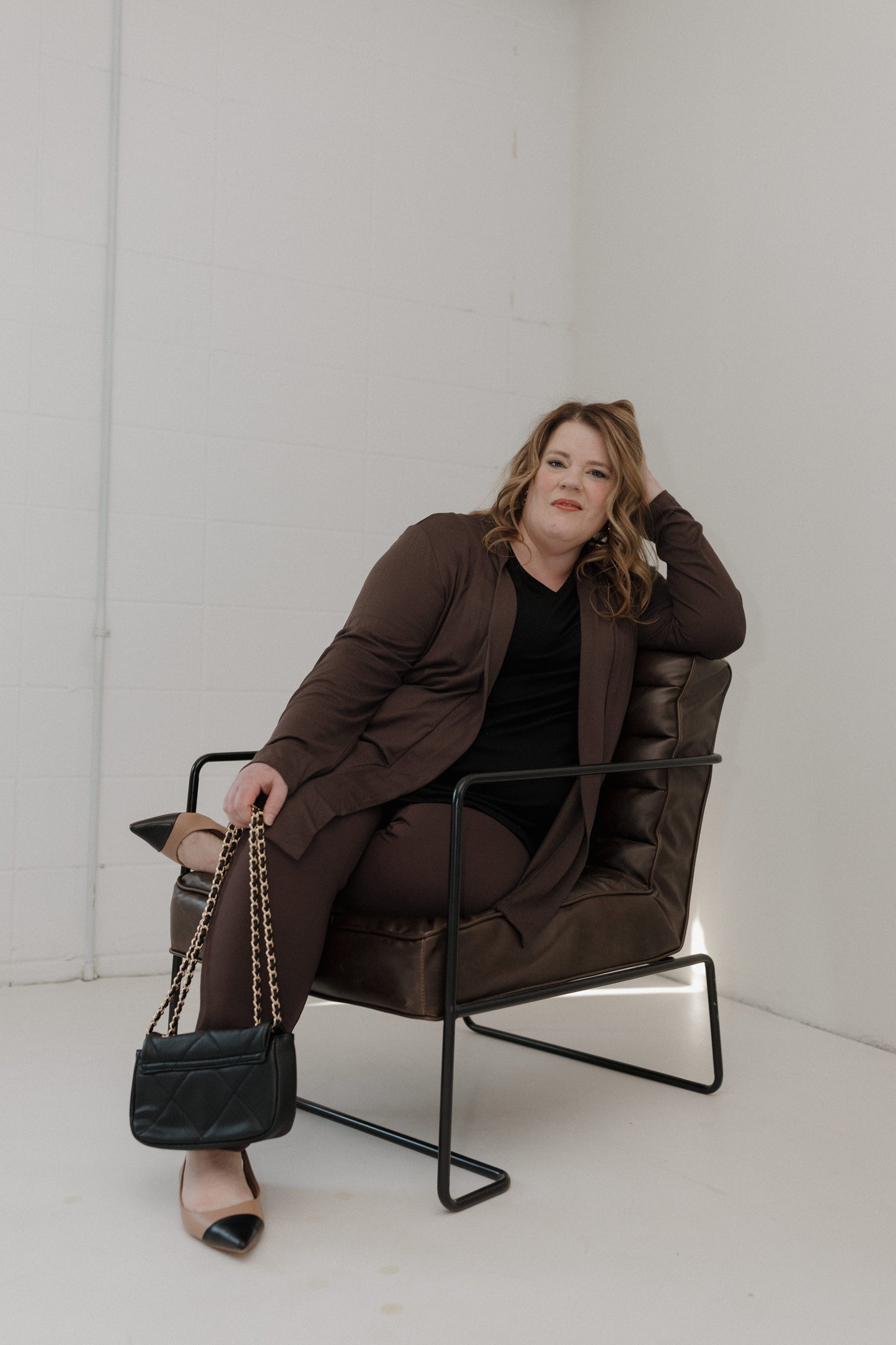 Woman sitting in a brown chair holding a black handbag against a white background