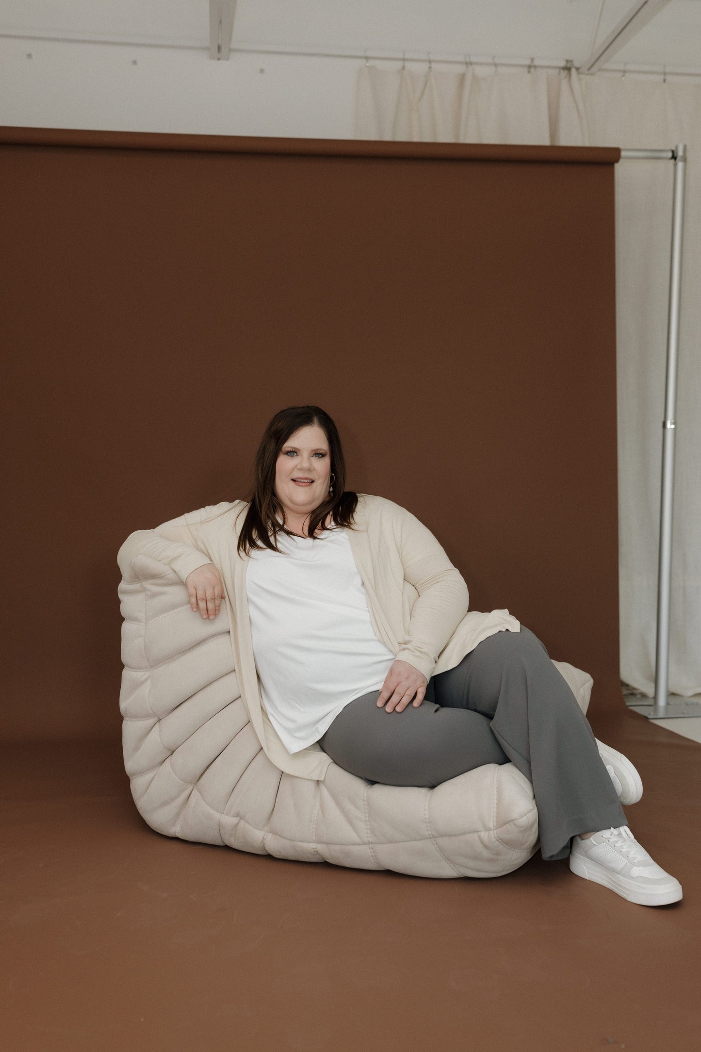 Woman sitting on a large beige bean bag chair against a brown wall.