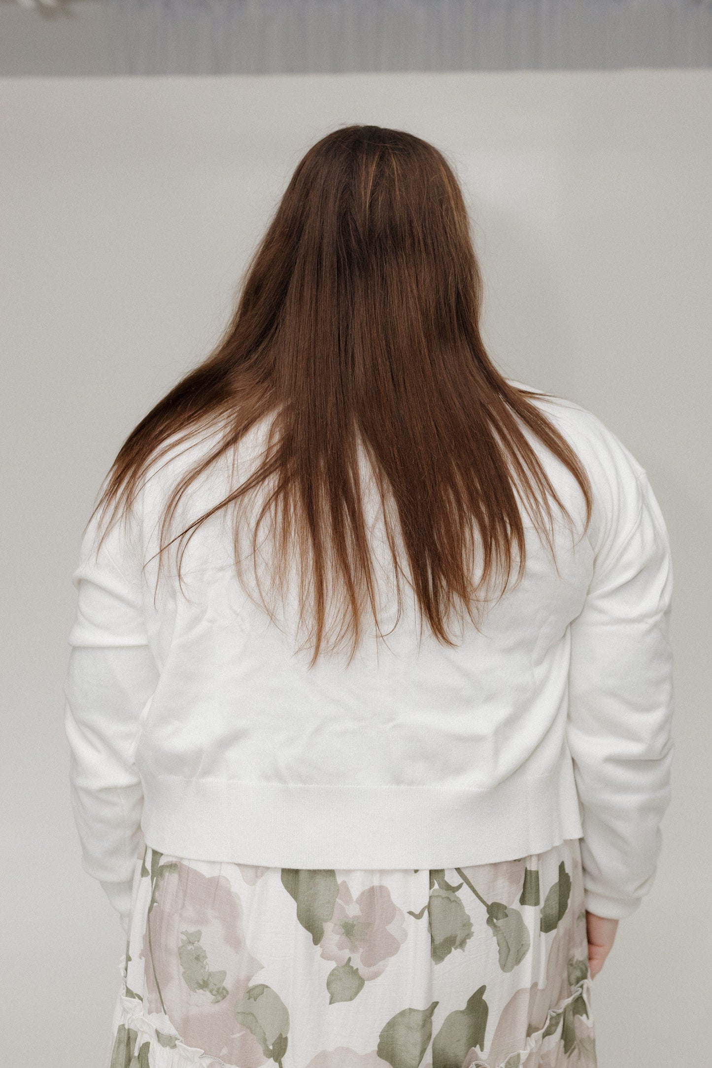 Person wearing a white sweater and floral skirt from behind against a plain background