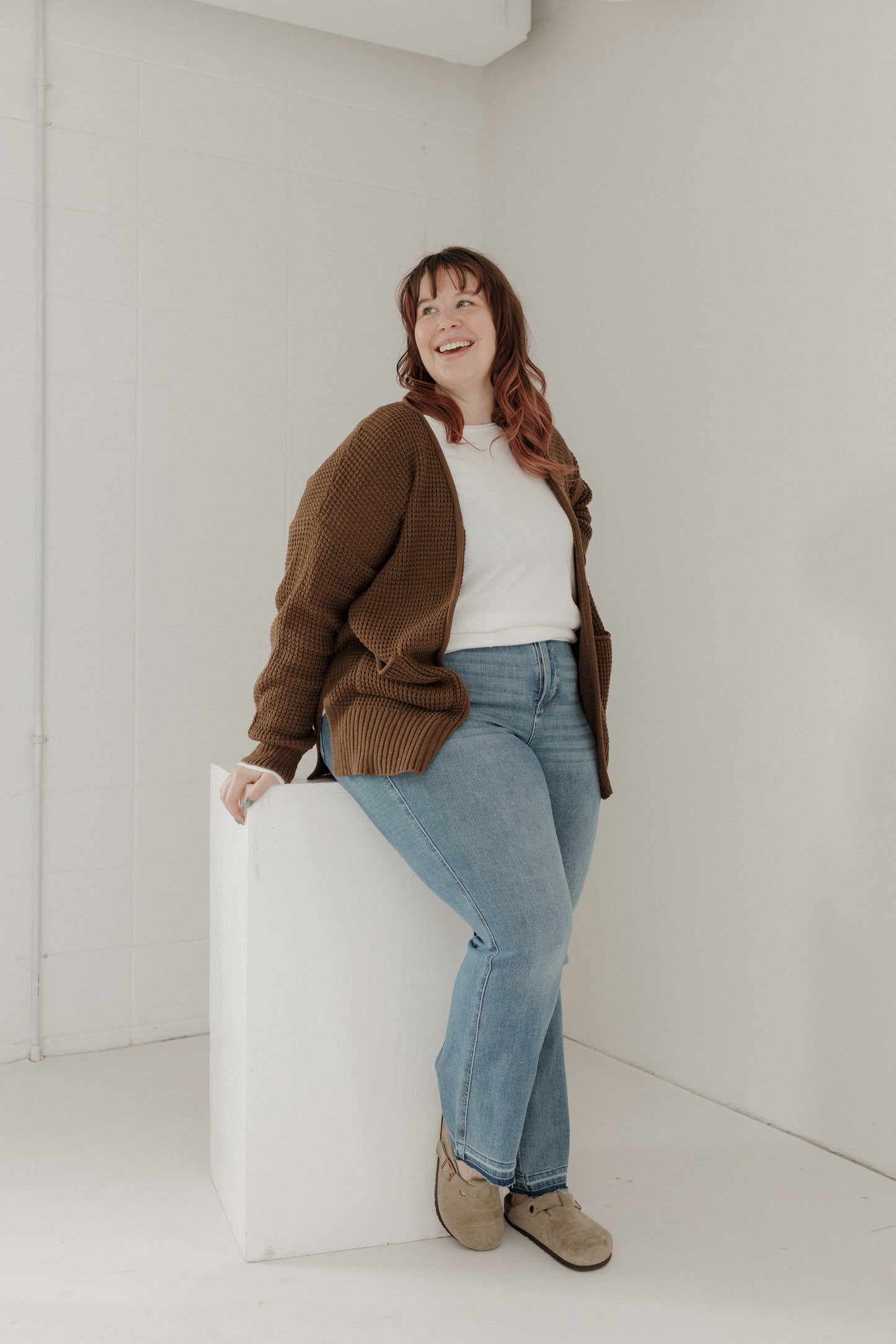 Woman sitting on a white cube wearing a brown cardigan, white shirt, blue jeans, and beige shoes in a minimalistic setting.
