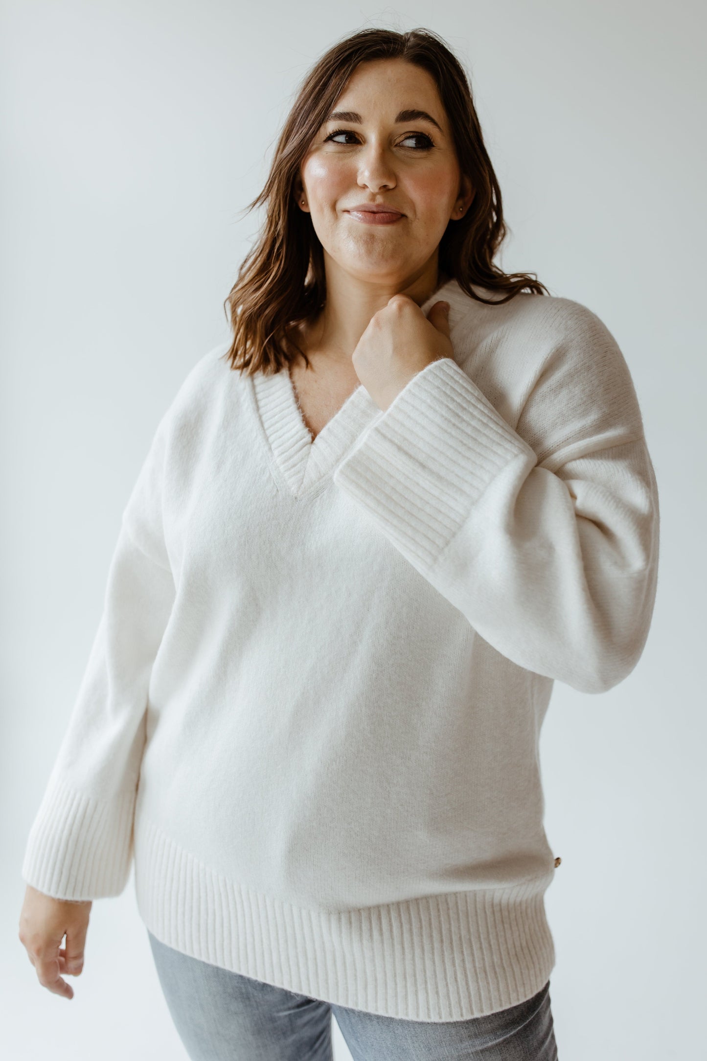 The woman stands gracefully against a plain background, showcasing a luxurious fabric in her white V-neck sweater paired effortlessly with jeans.