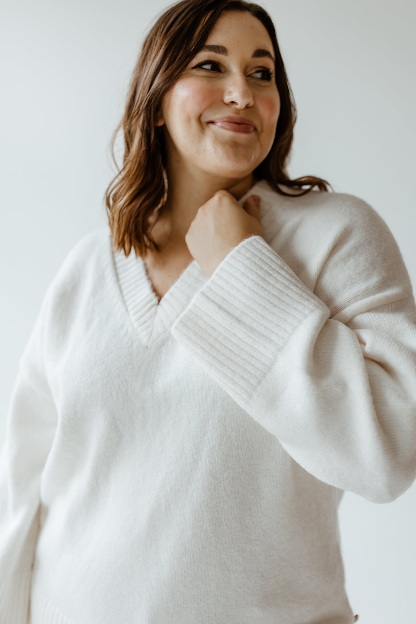 A woman in a luxurious V-neck sweater smiles and looks to the side, with one hand touching her neck.