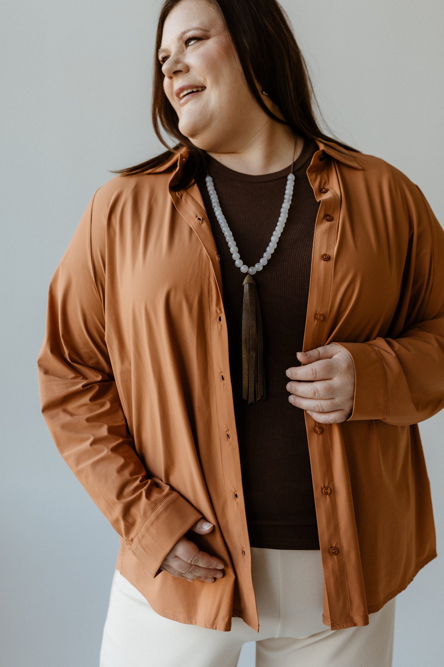 Person in a roasted cinnamon shirt with a large white bead necklace, smiling and looking to the side. The silky material adds an elegant touch, effortlessly blending comfort with style.