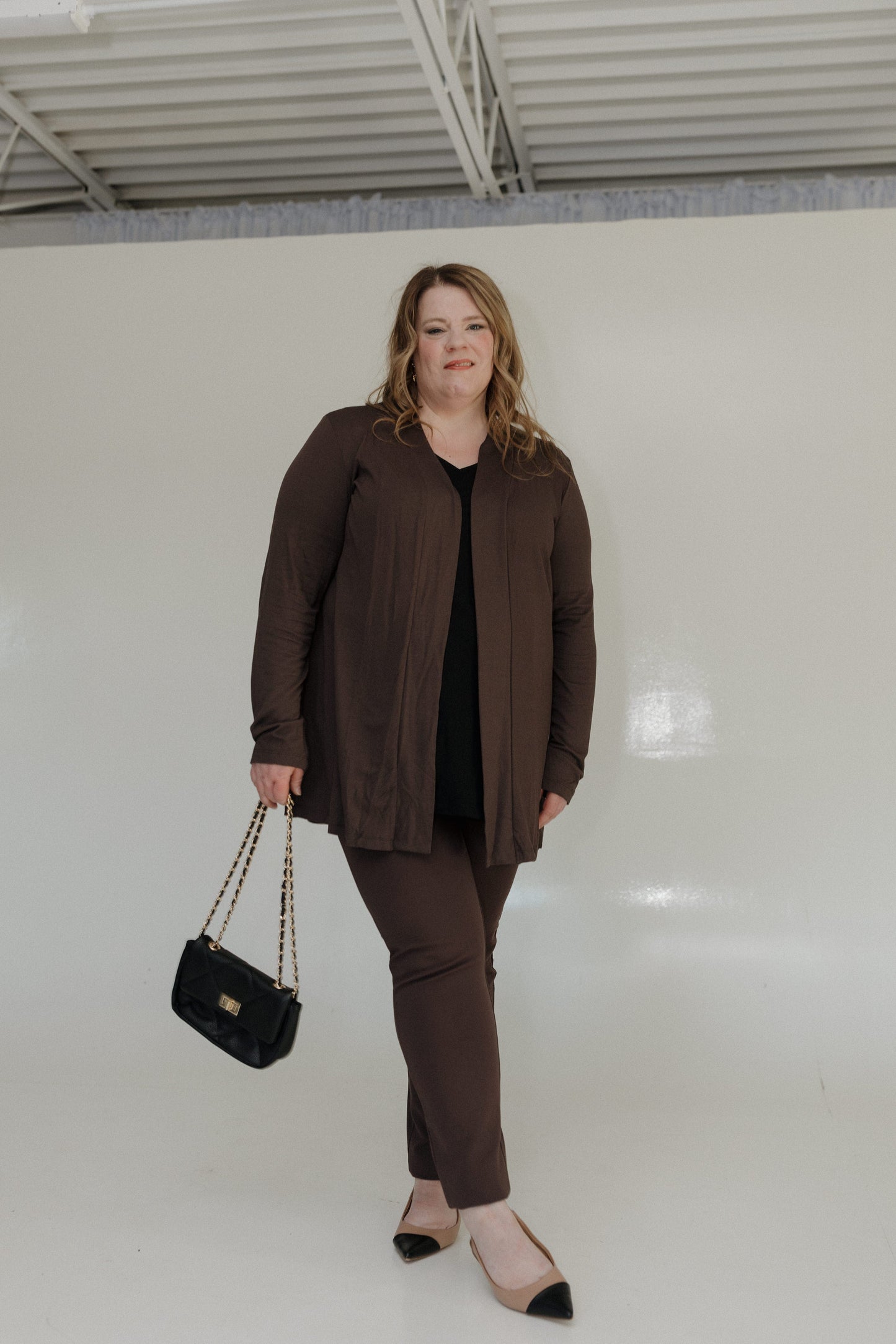 Woman in brown outfit holding a black handbag against a white wall