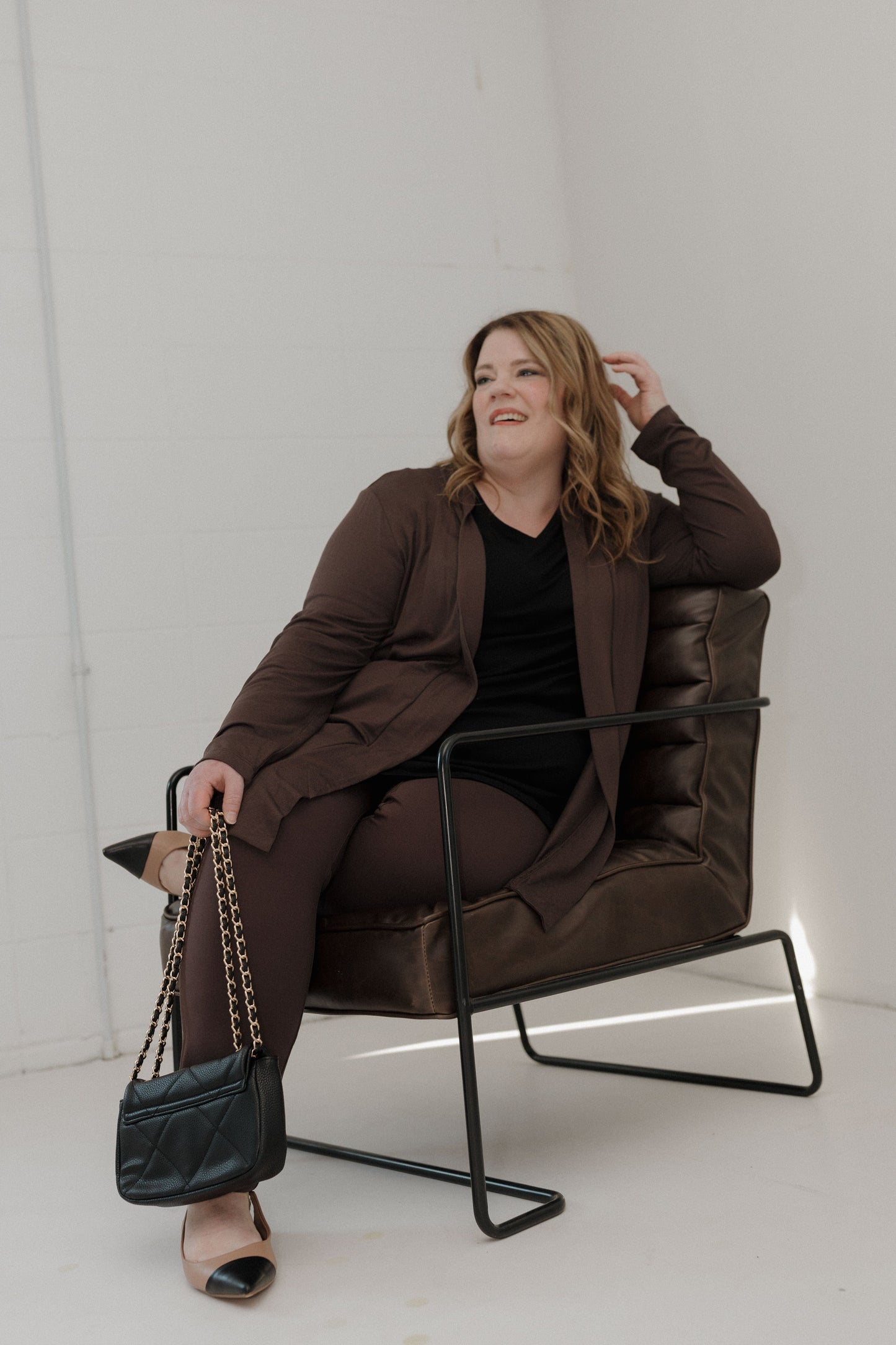 Woman sitting in a brown chair wearing a brown coat and black pants, holding a black handbag.