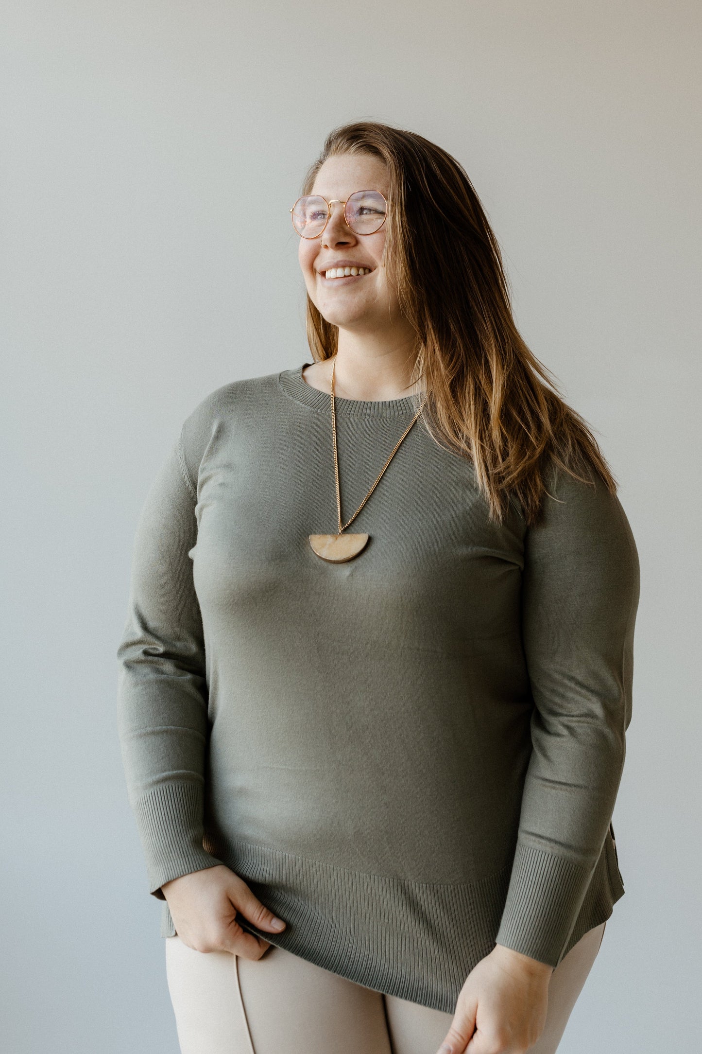 A smiling person wearing glasses and a luxurious knit green top with a necklace stands against a plain background.