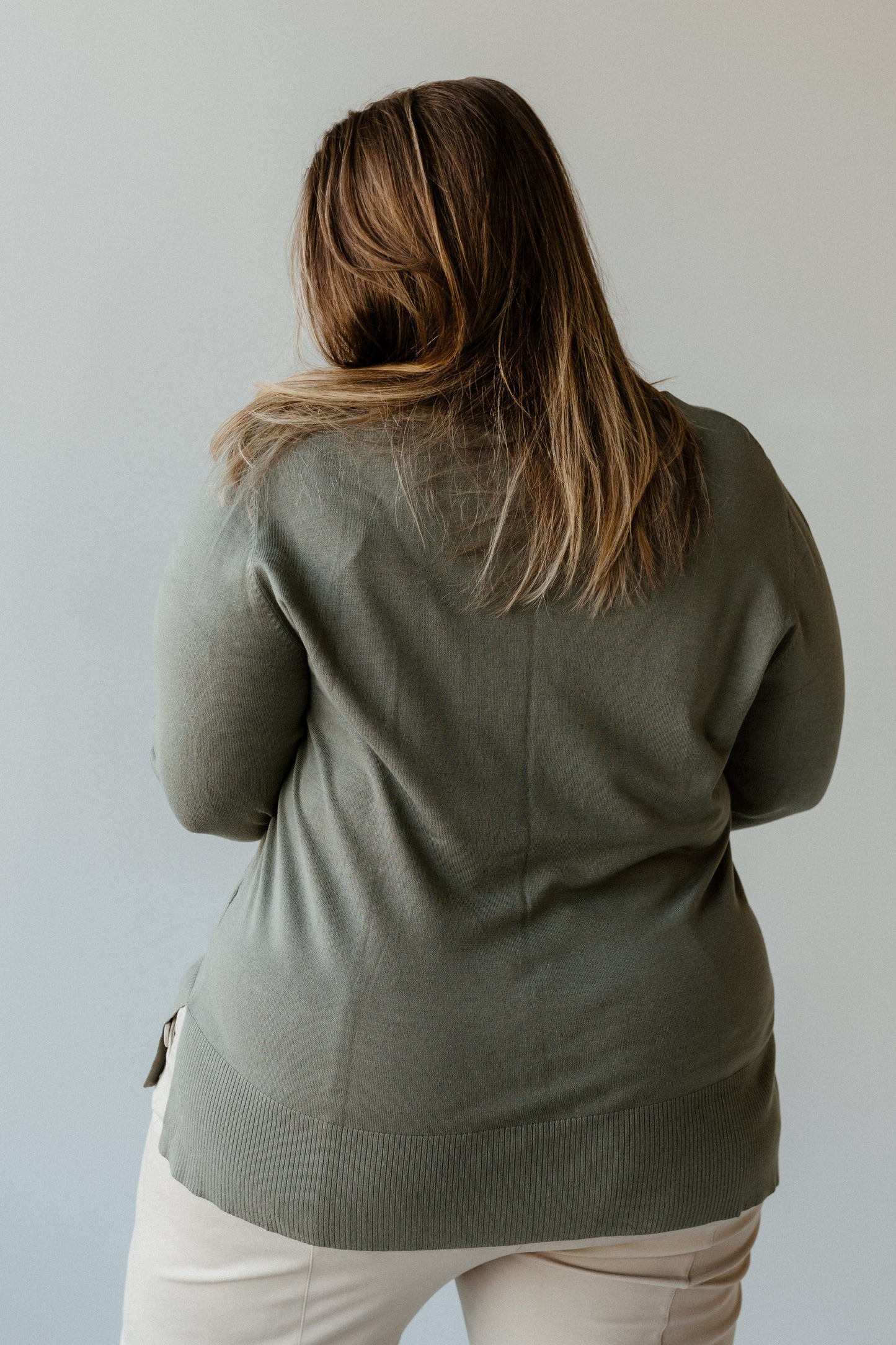 A person with long hair wearing a spiced rose lightweight tunic sweater and light-colored pants is standing, facing away from the camera.